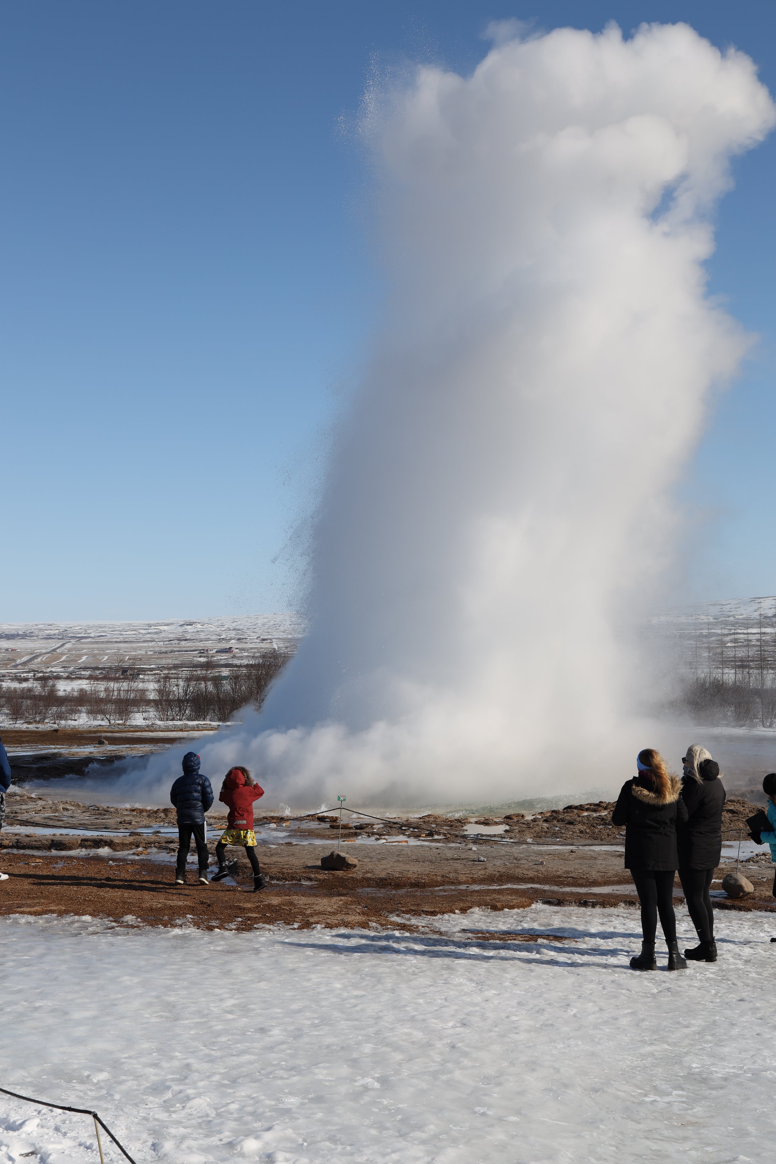 People observing a large geyser erupting in a cold, snowy landscape.
