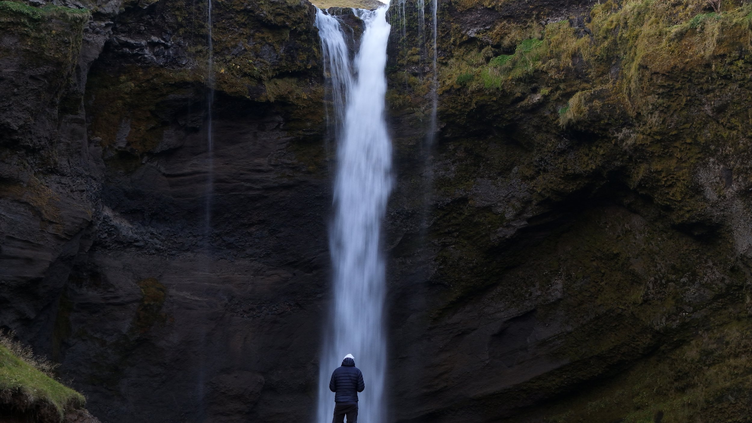 Person standing in front of a tall, narrow waterfall flowing down a rocky cliffside.