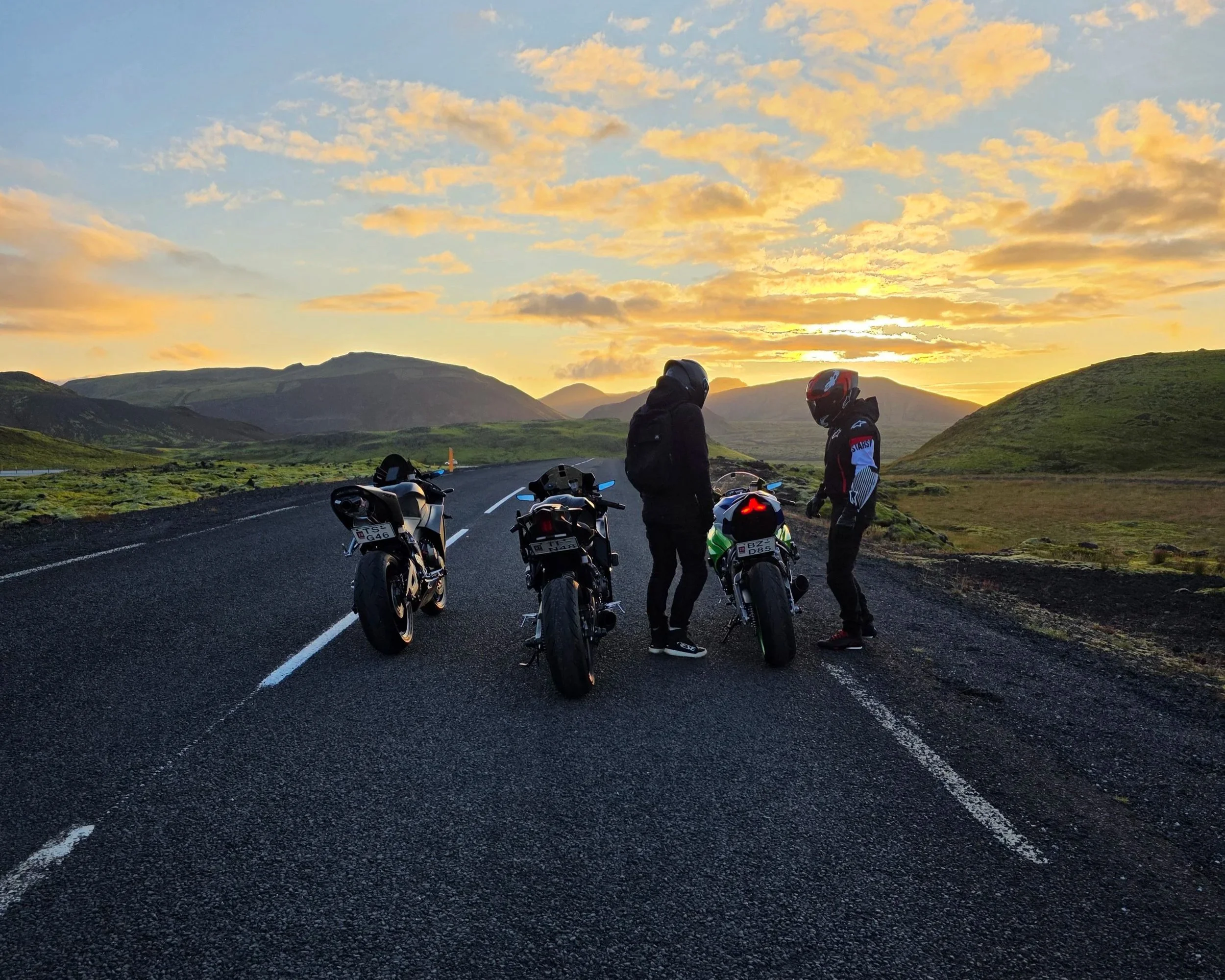 Two motorcyclists with helmets off are standing beside their bikes on a scenic mountain road at sunset, with three parked motorcycles nearby and lush green hills in the background.