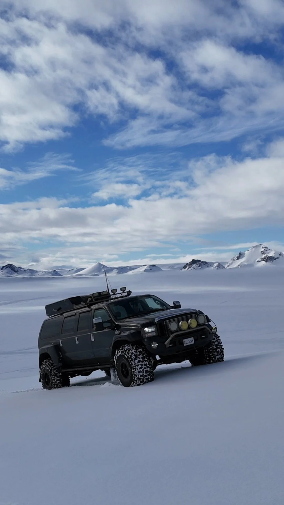 A black off-road vehicle parked on snow in a snowy landscape with mountains and a partly cloudy sky in the background.