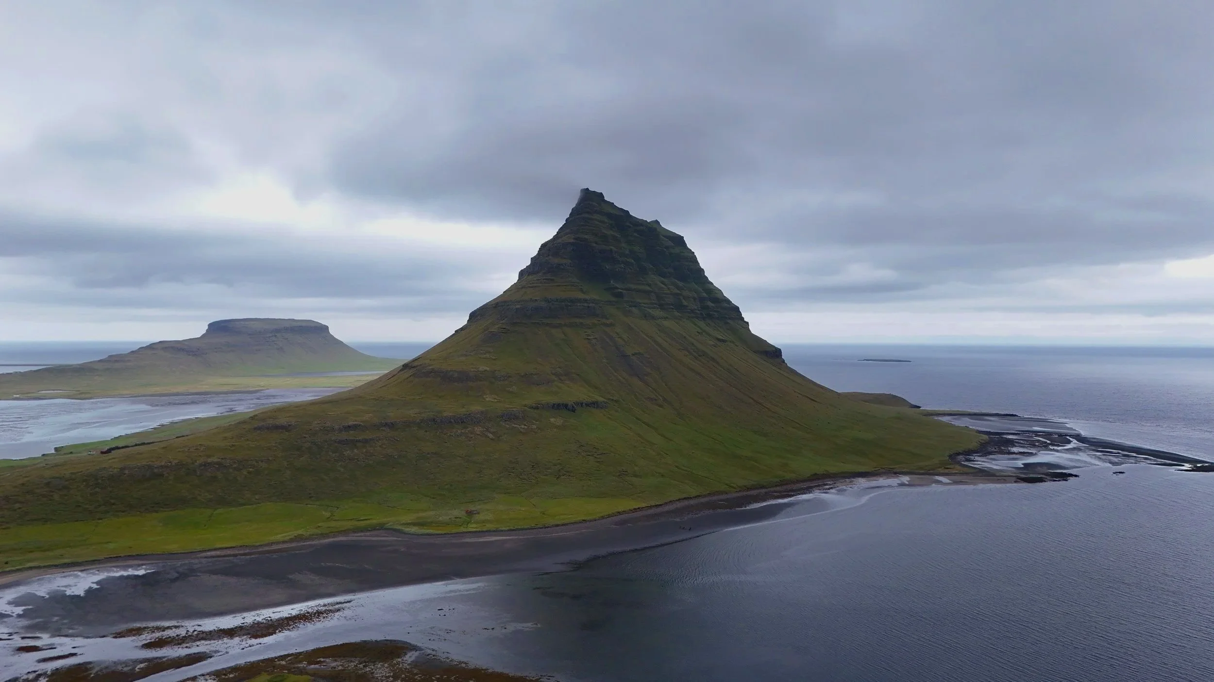 A scenic landscape featuring a prominent sculpted mountain with a broad base and pointed peak, surrounded by smaller hills, water bodies, and a cloudy sky.