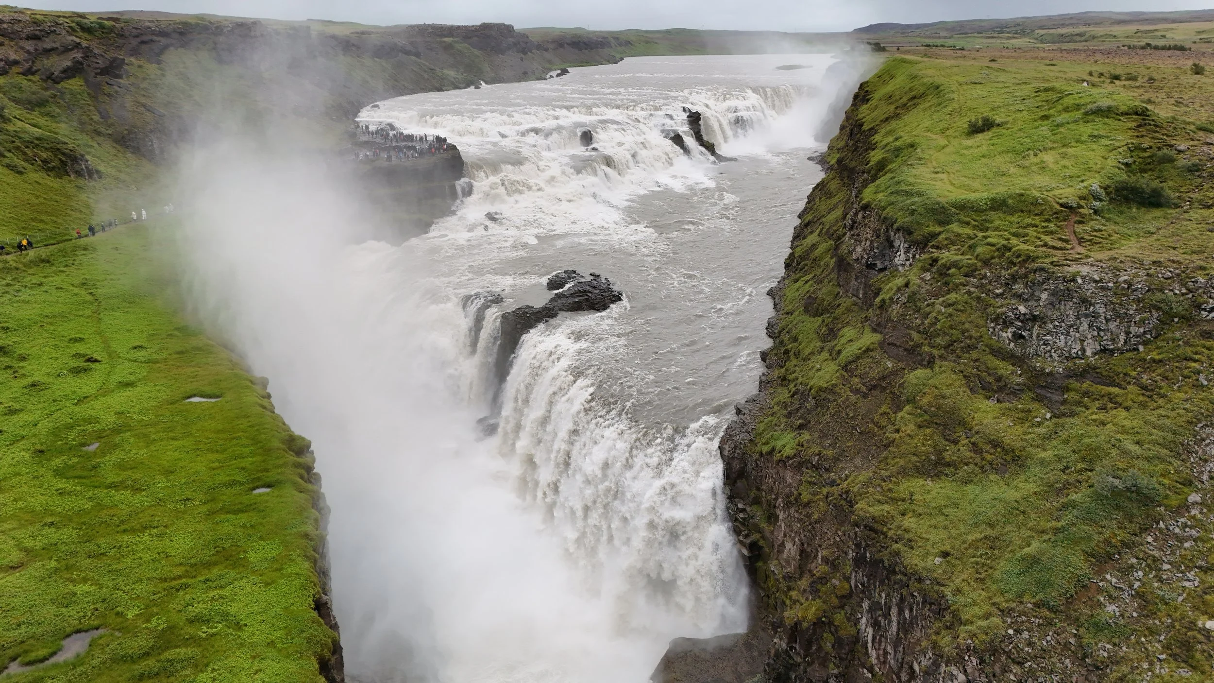 A large waterfall with mist rising from it, surrounded by green cliffs and grassy landscape, with a group of people walking along a path on the left.