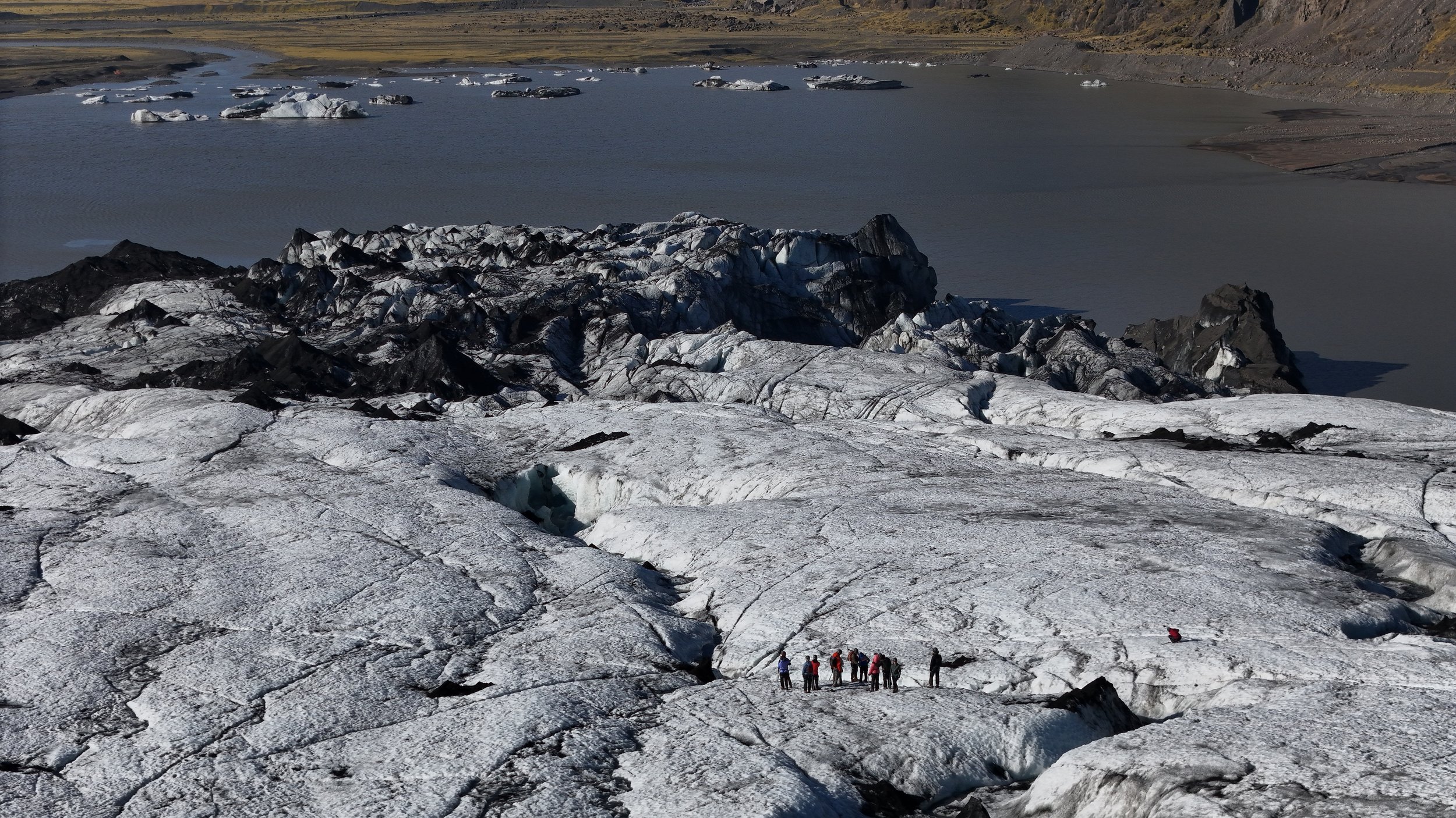 Group of hikers standing on a glacier with black and white ice, overlooking a lake with floating ice chunks, in a mountainous landscape.