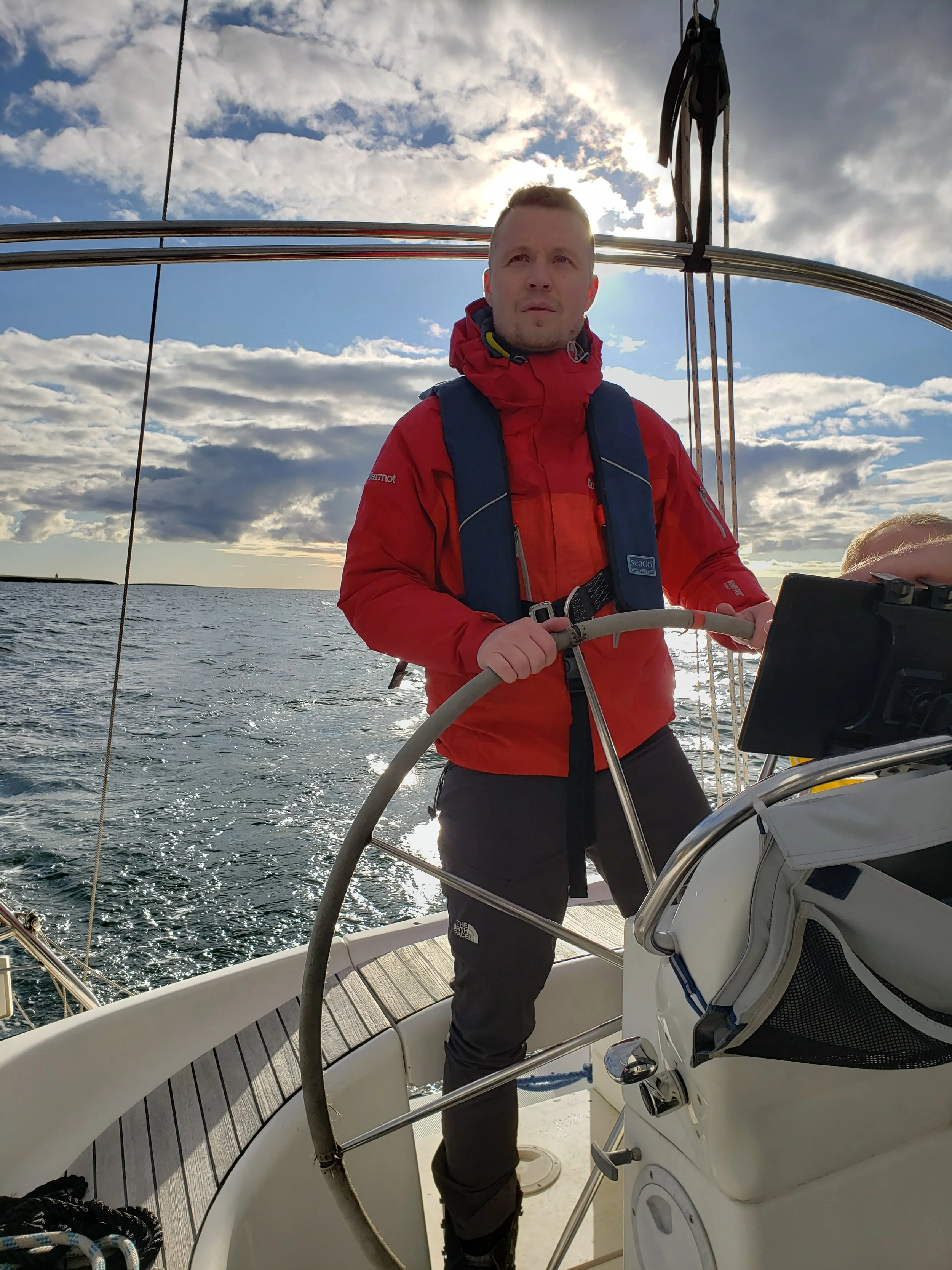 A man in a red jacket and black pants standing at the helm of a sailboat, holding the steering wheel with a body of water and a cloudy sky in the background.