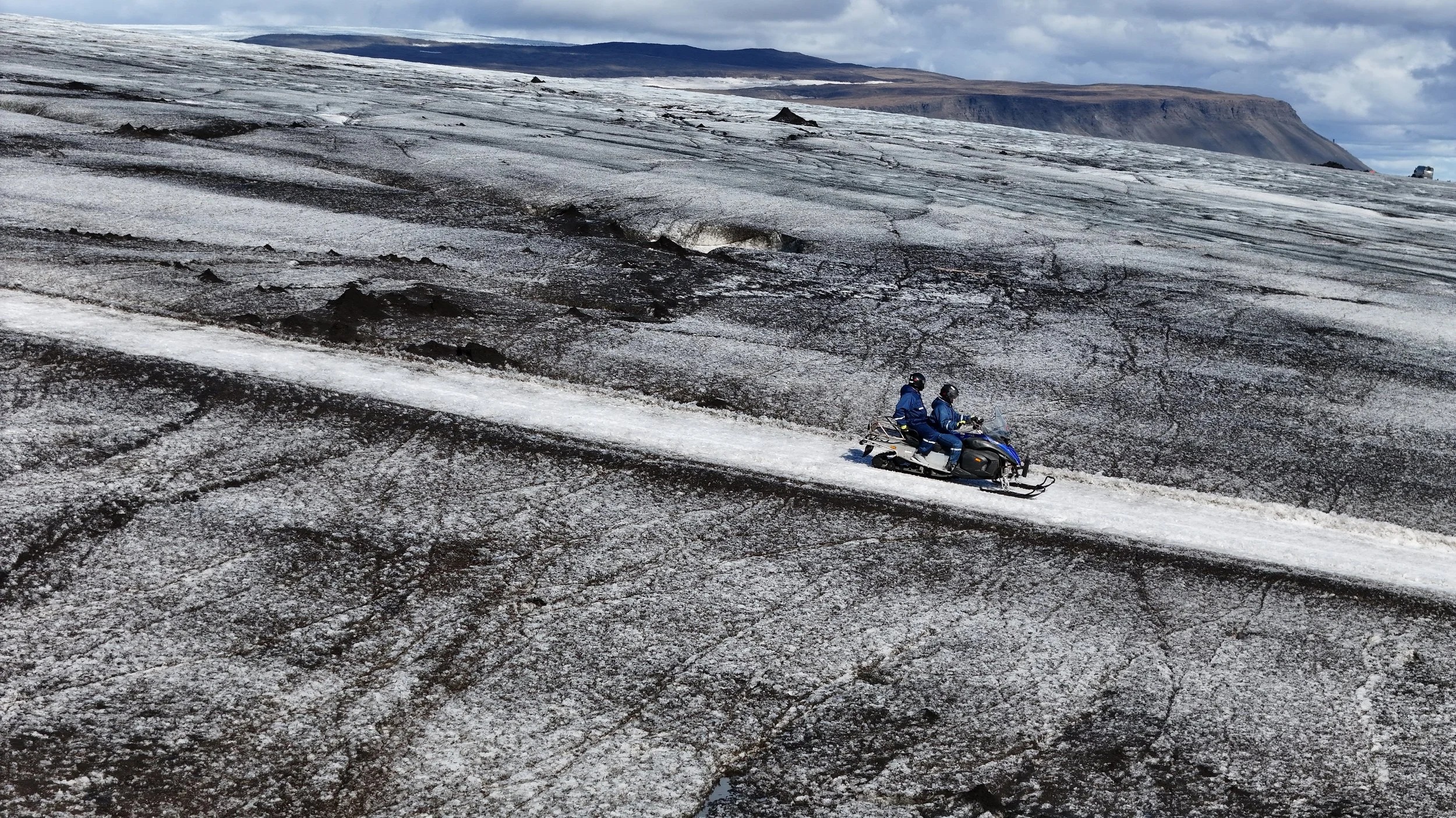 Two people riding a snowmobile on a snowy, rocky landscape with mountains in the background.
