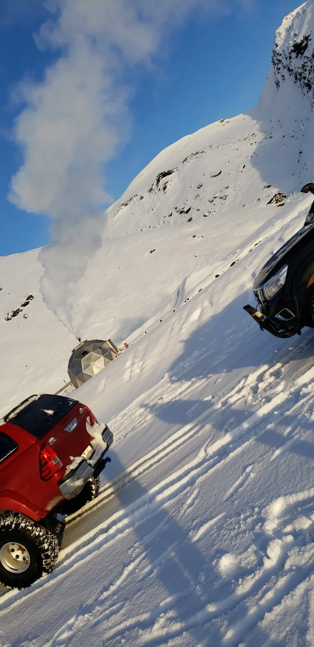 Snow-covered mountain landscape with a geodesic dome and parked cars, some snowfall on their surfaces, and a plume of smoke or steam coming from the dome, under a partly cloudy sky.