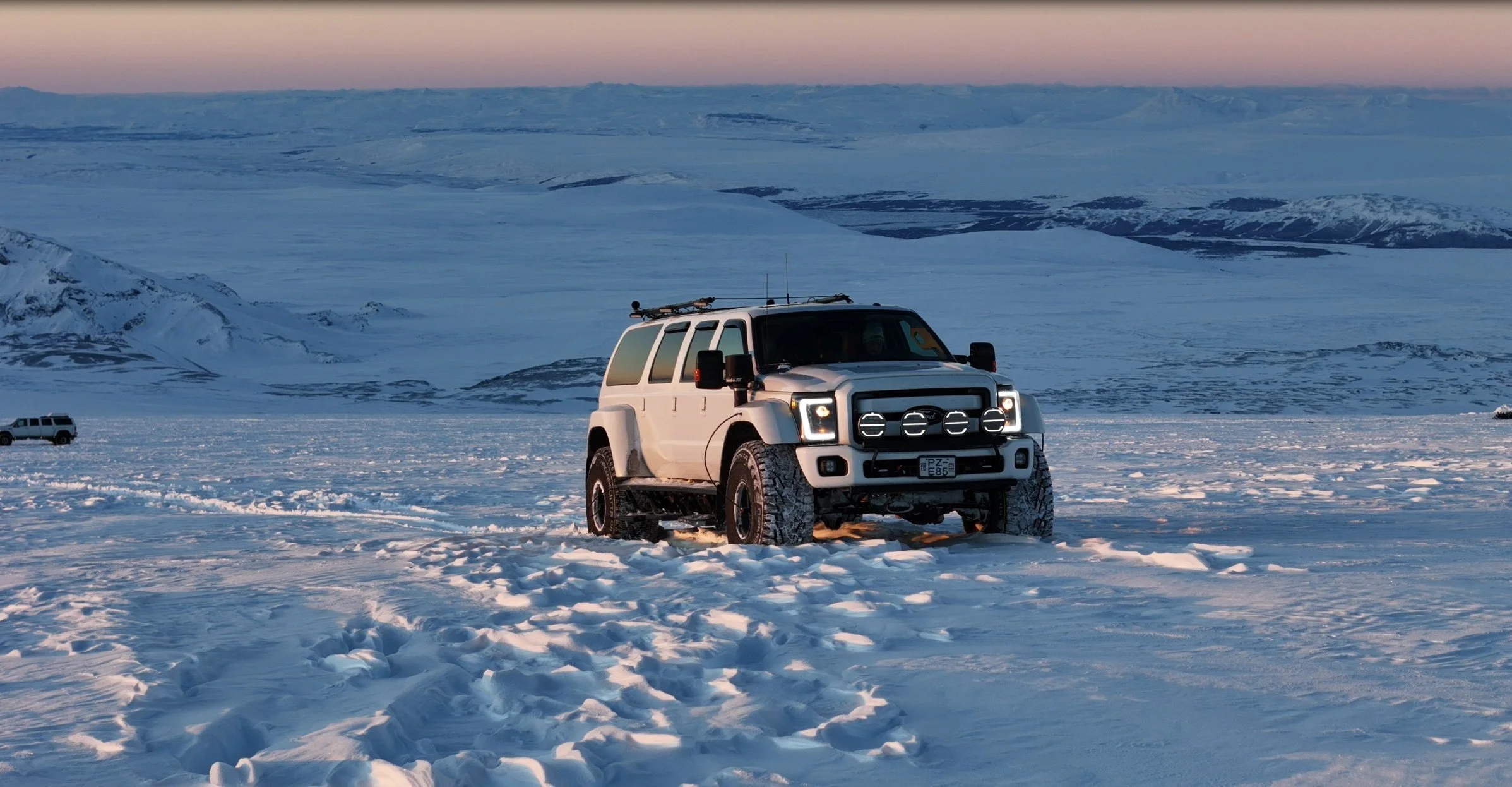 A white off-road vehicle driving in the snow-covered landscape during sunset, with mountains in the background.