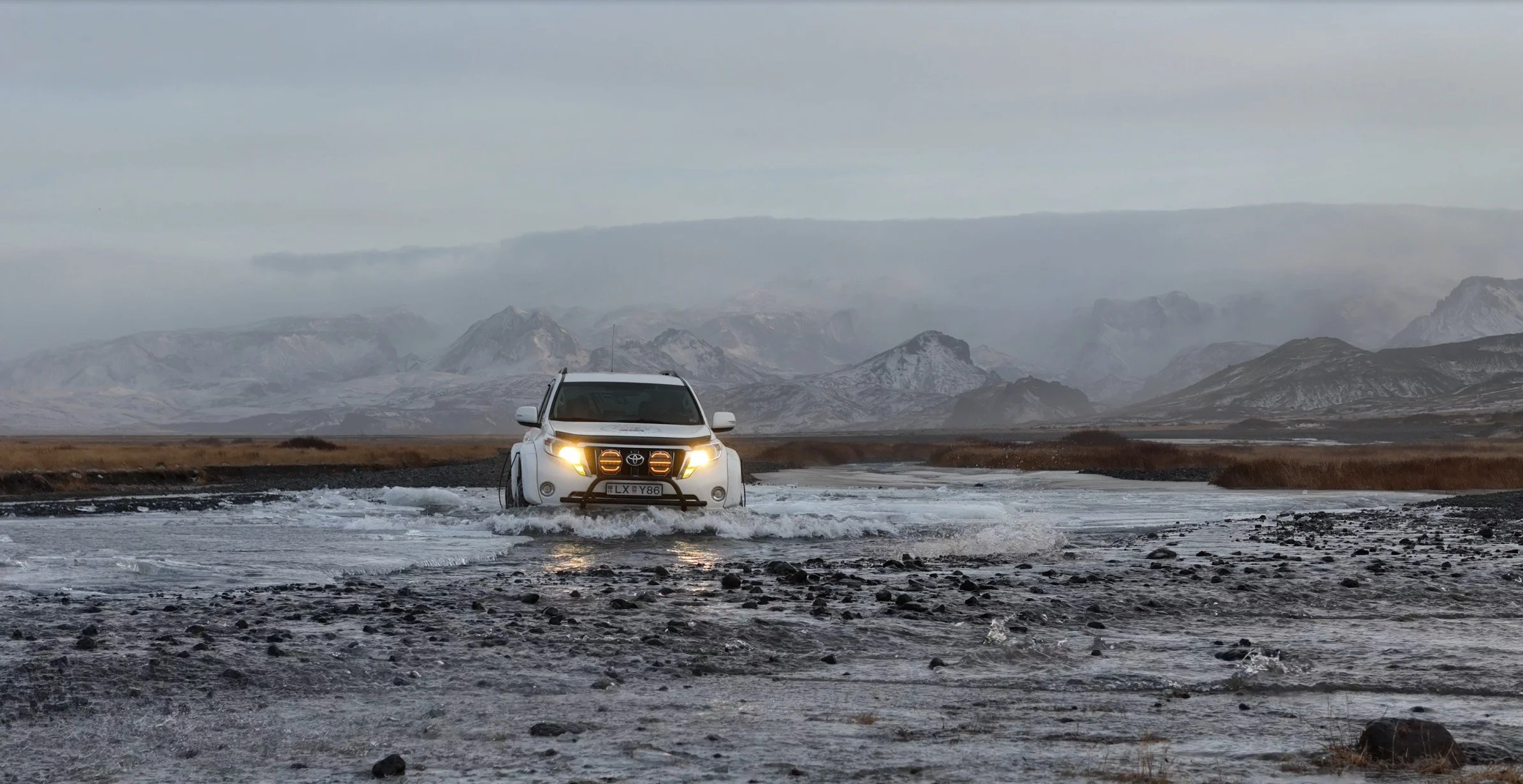 White SUV driving through a shallow river in a rugged landscape with mountains in the background