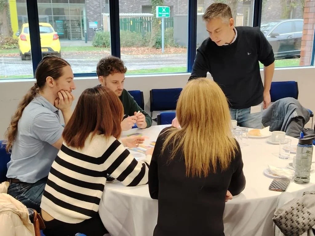 A group of five people, gathered around a table in a meeting room with large windows showing cars and a street outside. The man -Rich Allen- appears to be explaining something to them.