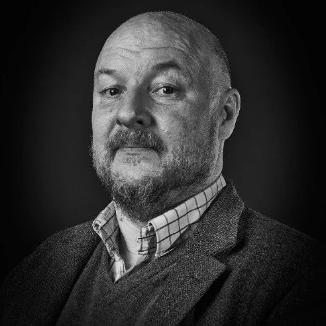 Black and white portrait of Simon Wardley with a beard and short hair, wearing a checked shirt and a blazer, looking at the camera.