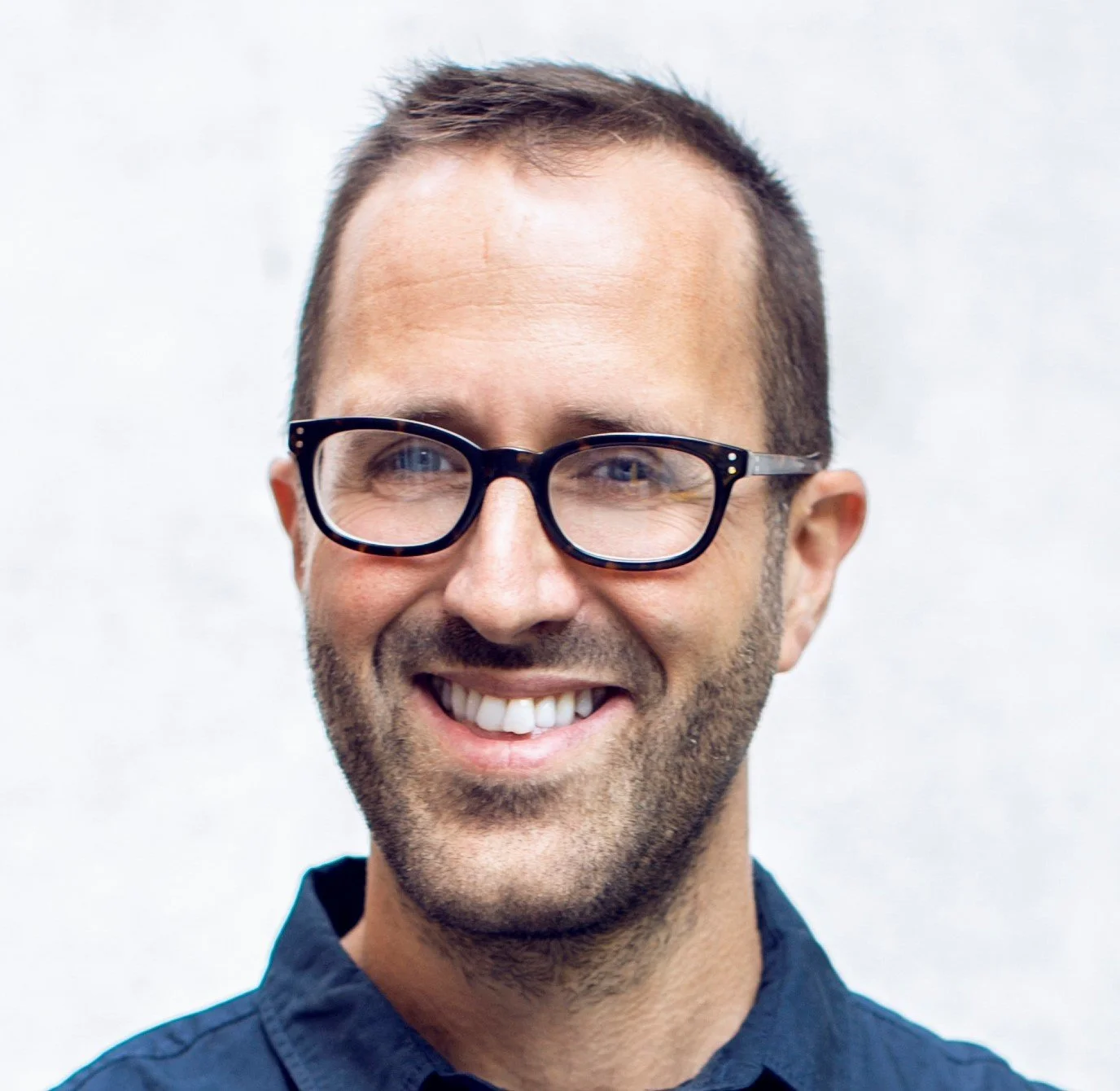 A smiling man, Tom Quay, with glasses and a dark blue shirt against a plain white background.