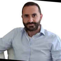 Portrait of a man with a beard, wearing a light blue button-up shirt, smiling lightly, against a white background.