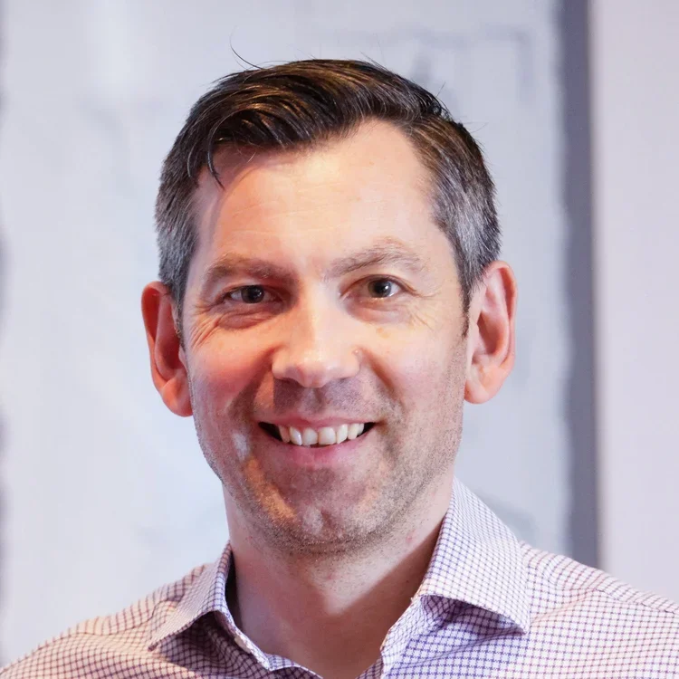 A smiling man, Matthew Skelton, with short dark hair, wearing a checkered shirt, standing indoors against a blurred background.