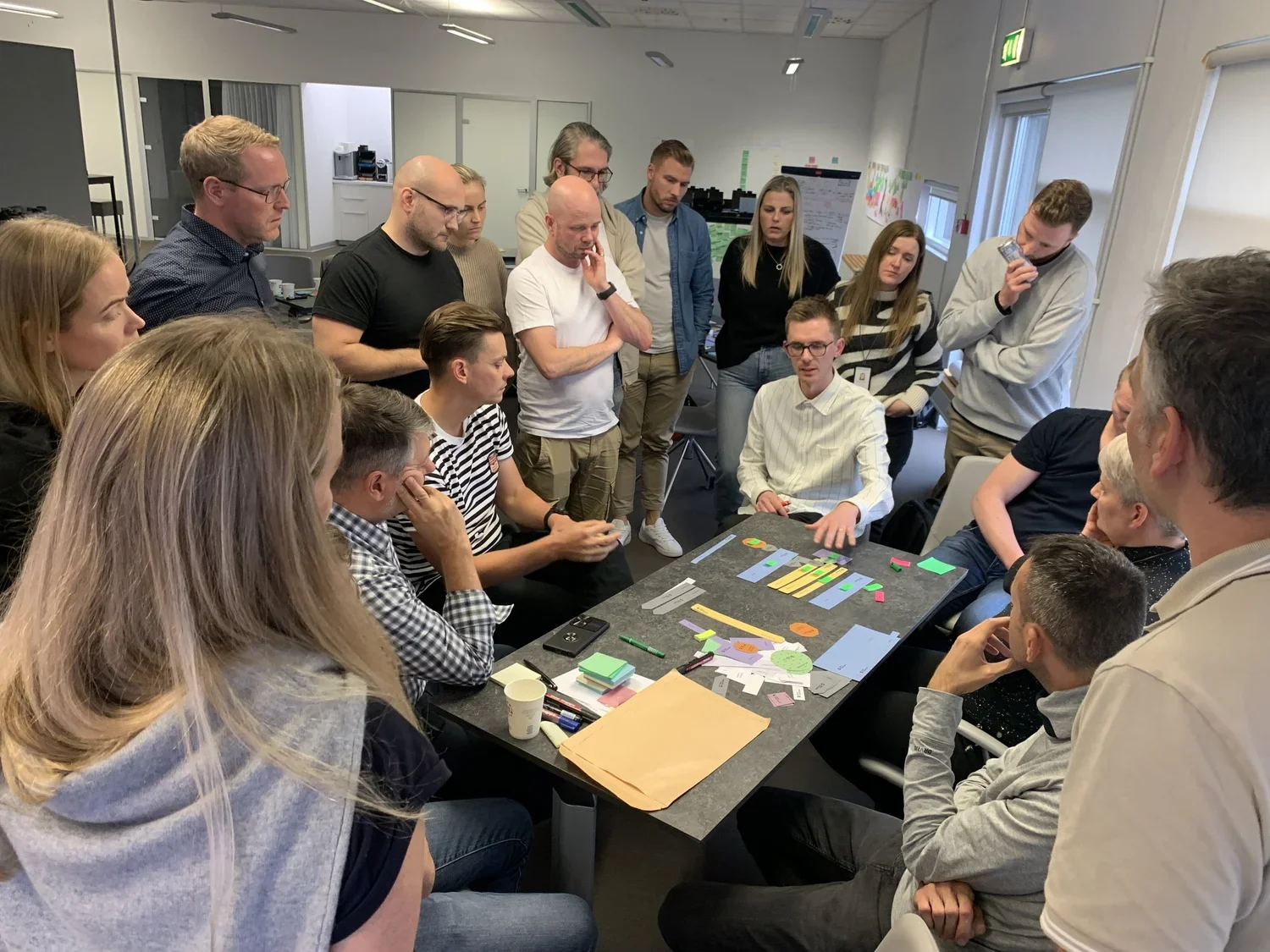 Group of people gathered around a table in a meeting room, listening to a woman speaking. The table has colorful paper cutouts and markers, indicating a collaborative activity or brainstorming session.