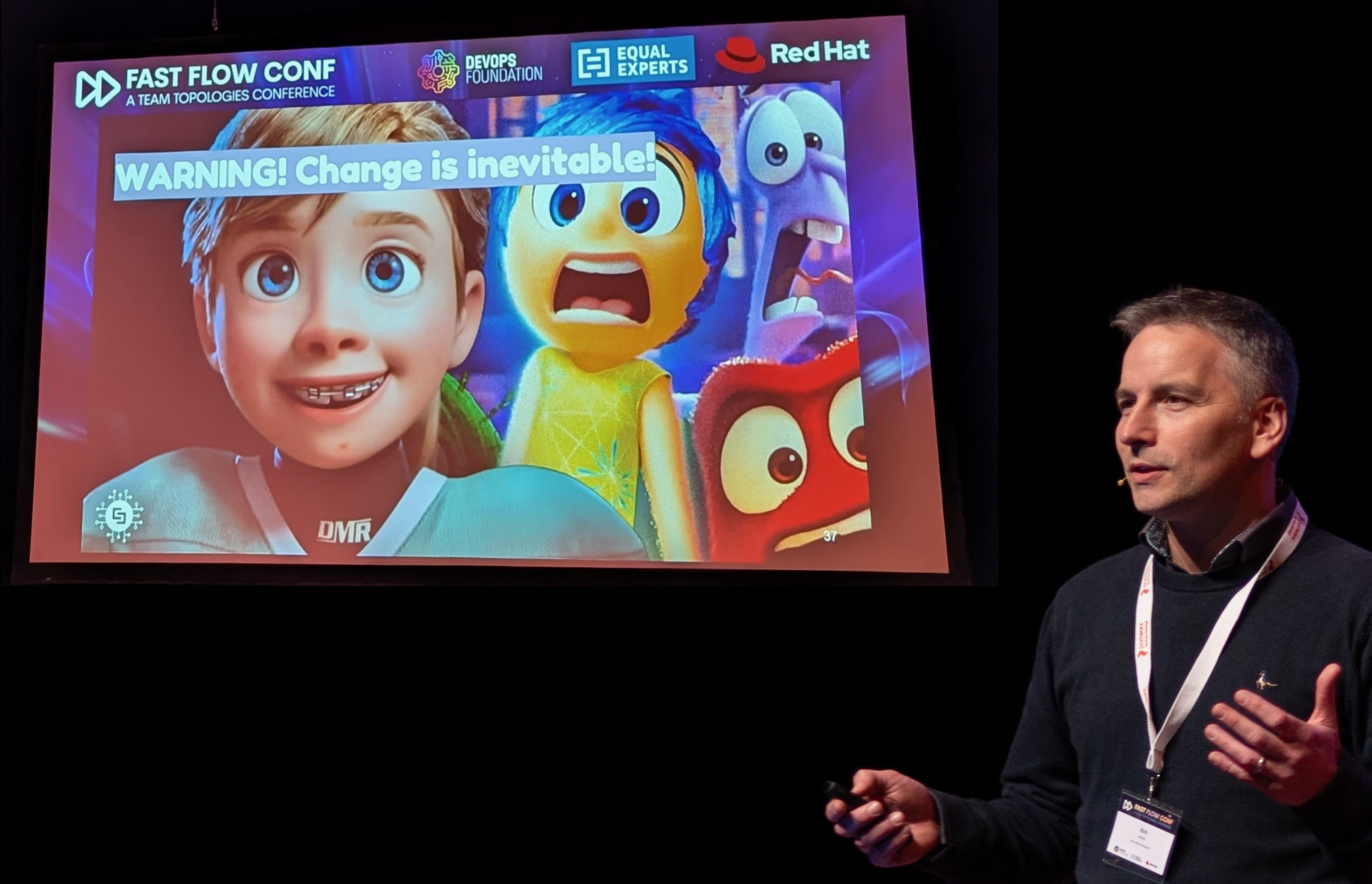 Rich Allen gives a presentation at a conference with a slide showing animated characters. The slide displays a warning message "WARNING! Change is inevitable!".