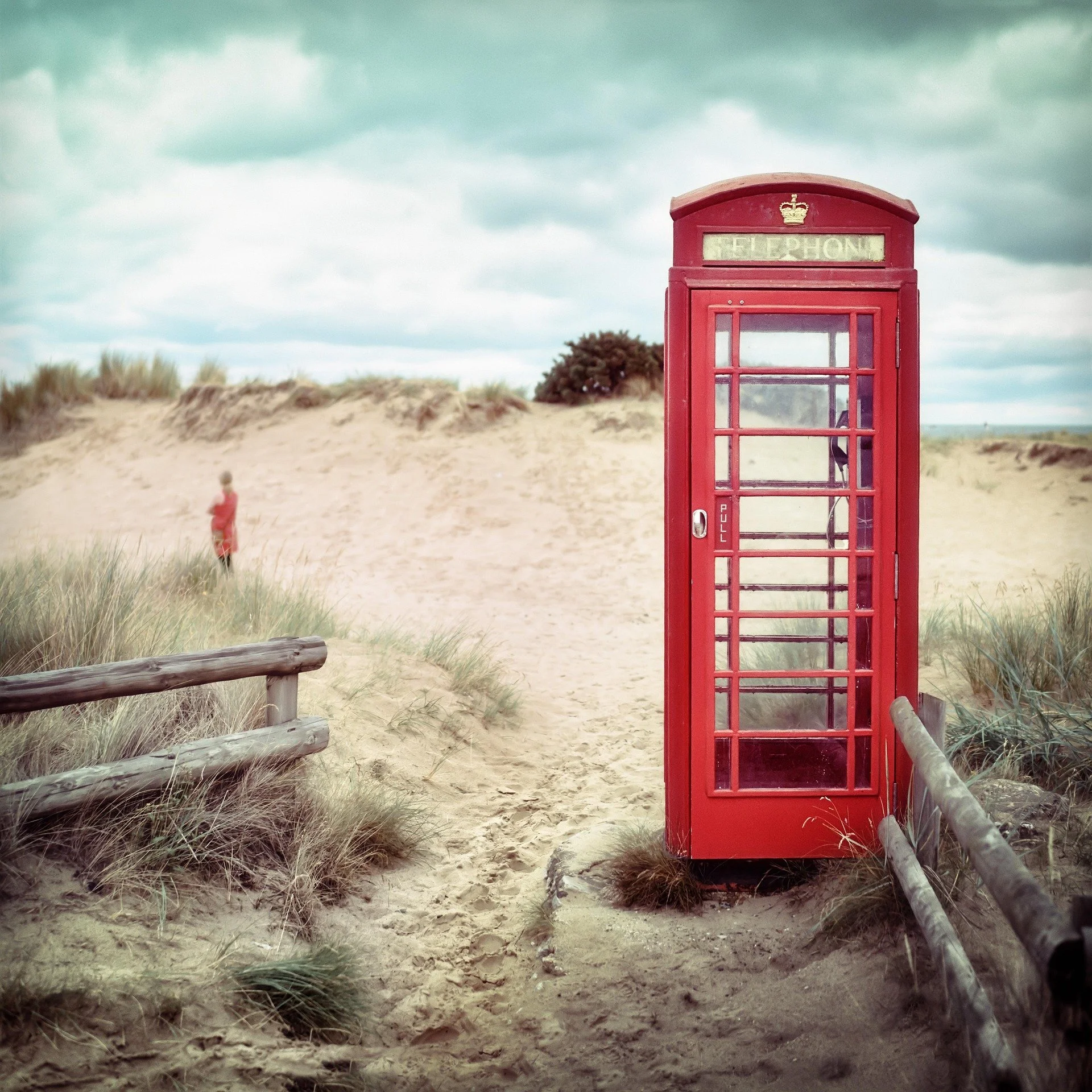 A red British telephone booth on sandy dunes with a cloudy sky in the background. A person in a red shirt is walking in the distance.
