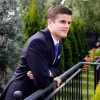 A young man in a dark suit and blue tie leaning on a black metal railing outdoors, with greenery and trees in the background.