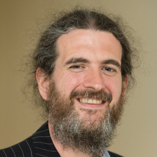 A smiling man with long hair and a beard, wearing a black suit with thin white pinstripes.