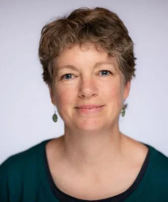 A woman with short, curly light brown hair and blue eyes, wearing a teal top with black trim and green earrings, smiling against a light gray background.