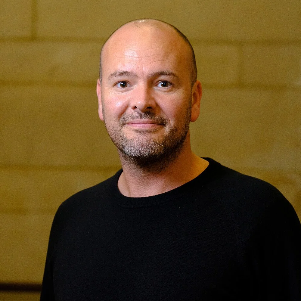 A man with a shaved head and short beard wearing a black shirt, standing against a wooden wall background.