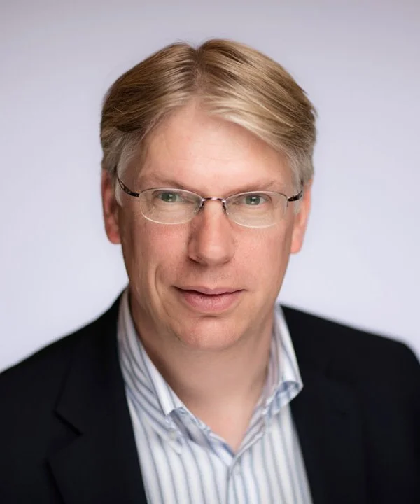 A professional headshot of a man with blonde hair and glasses, wearing a dark blazer and a light striped shirt against a plain background.