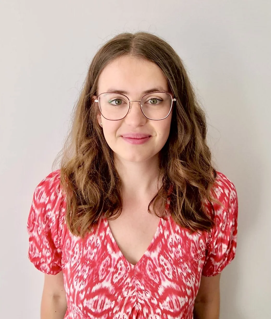 A young woman with wavy brown hair, wearing glasses, a pink and white patterned top, stands against a plain light-colored background.