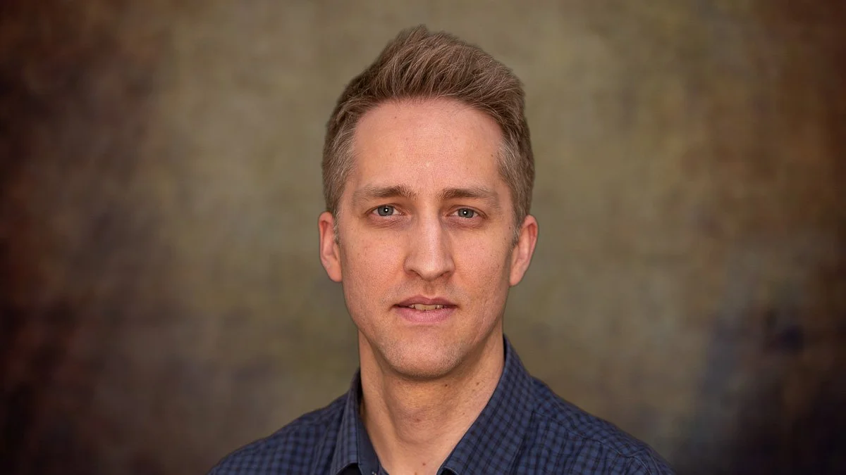 A professional portrait of a man with short light brown hair, wearing a dark blue and black checkered shirt, against a mottled brown background.