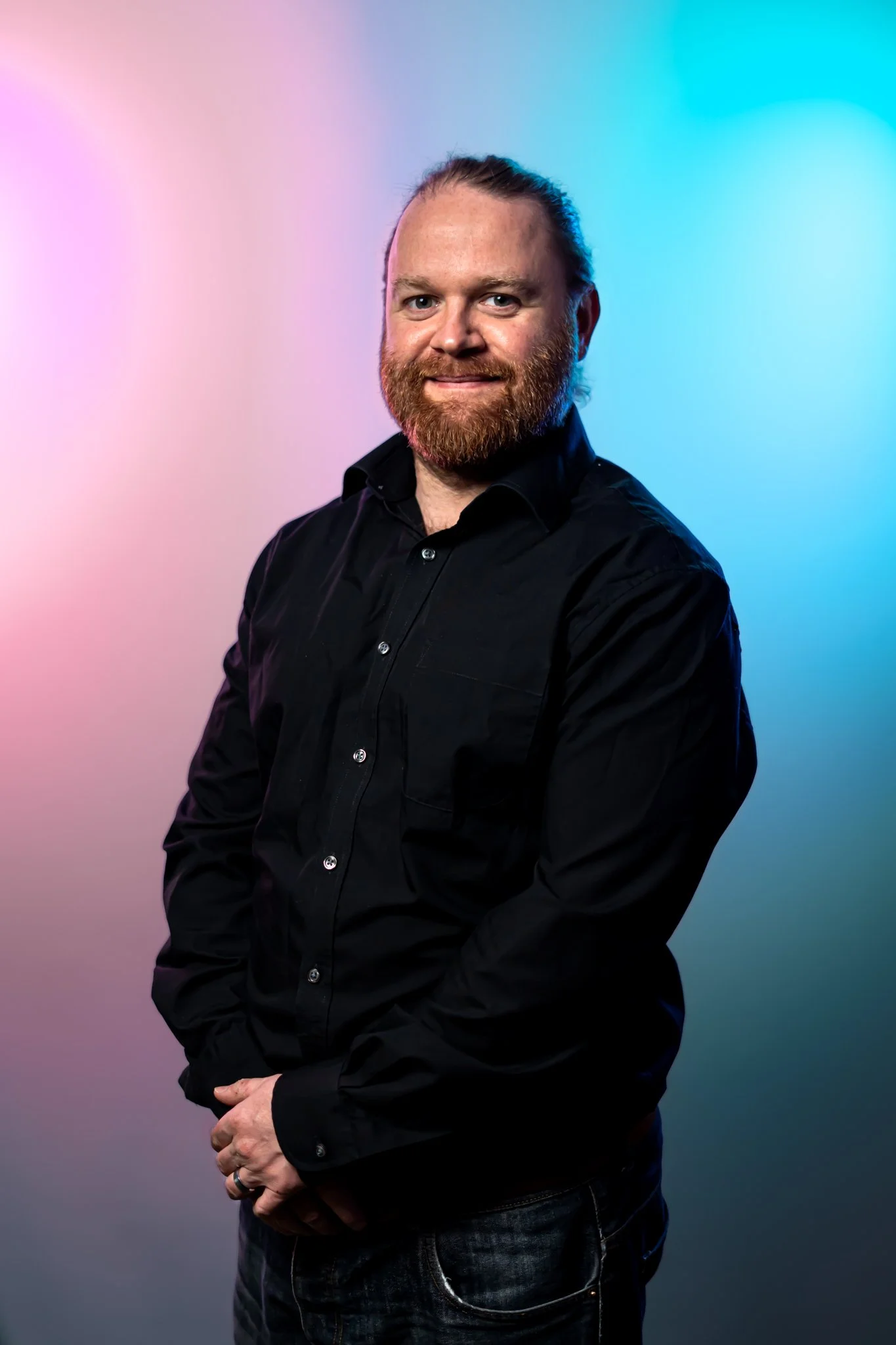 A man with a beard and long hair tied back, wearing a black button-up shirt, standing in front of a colorful gradient background with pink, purple, and blue hues.