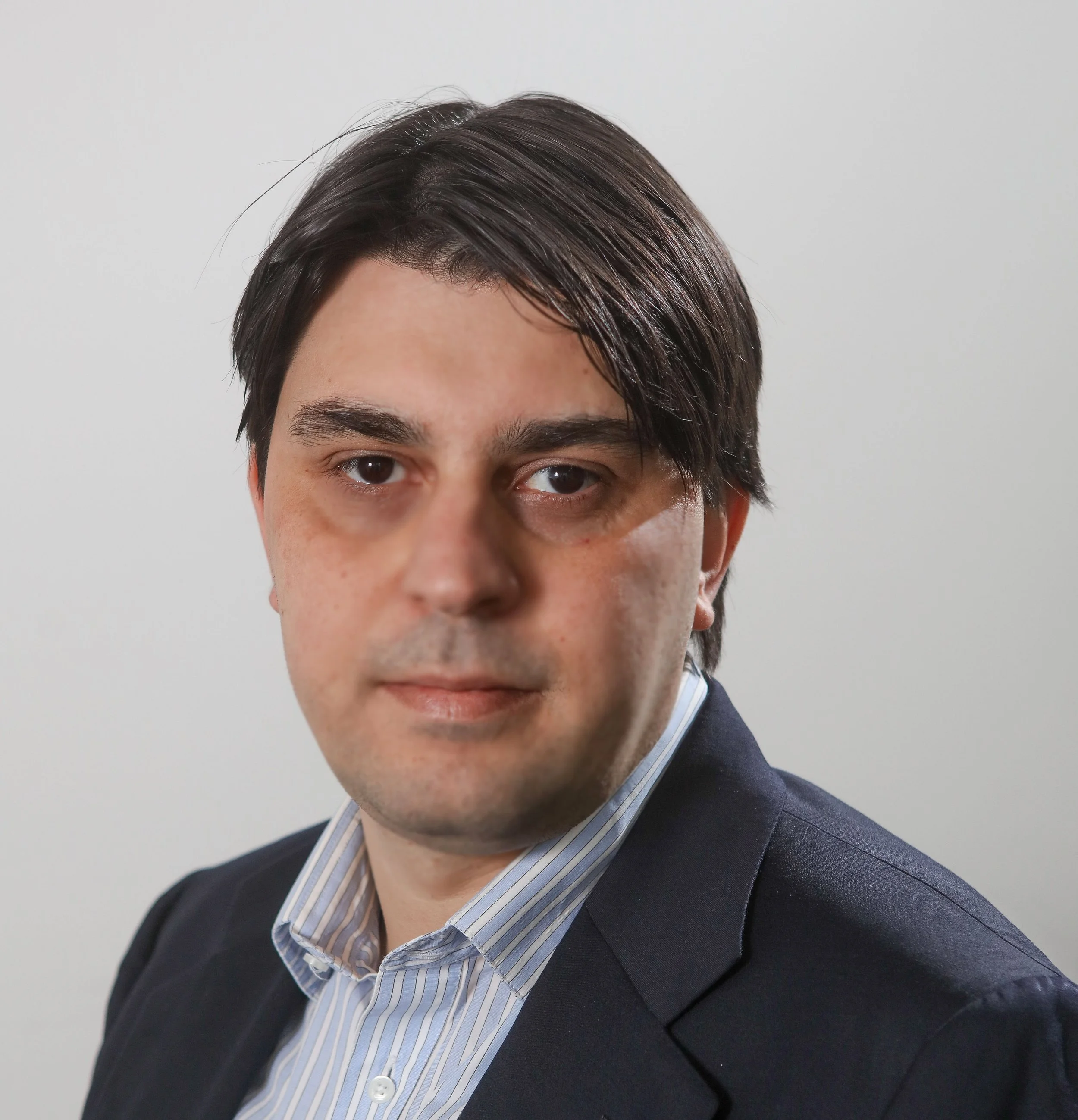 A professional headshot of a man with dark brown hair, wearing a dark suit jacket and a light blue striped shirt, against a plain white background.