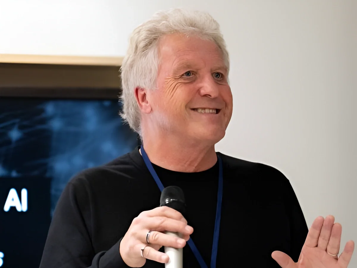 A smiling man with gray hair holding a microphone and making a friendly gesture during a presentation or speech.
