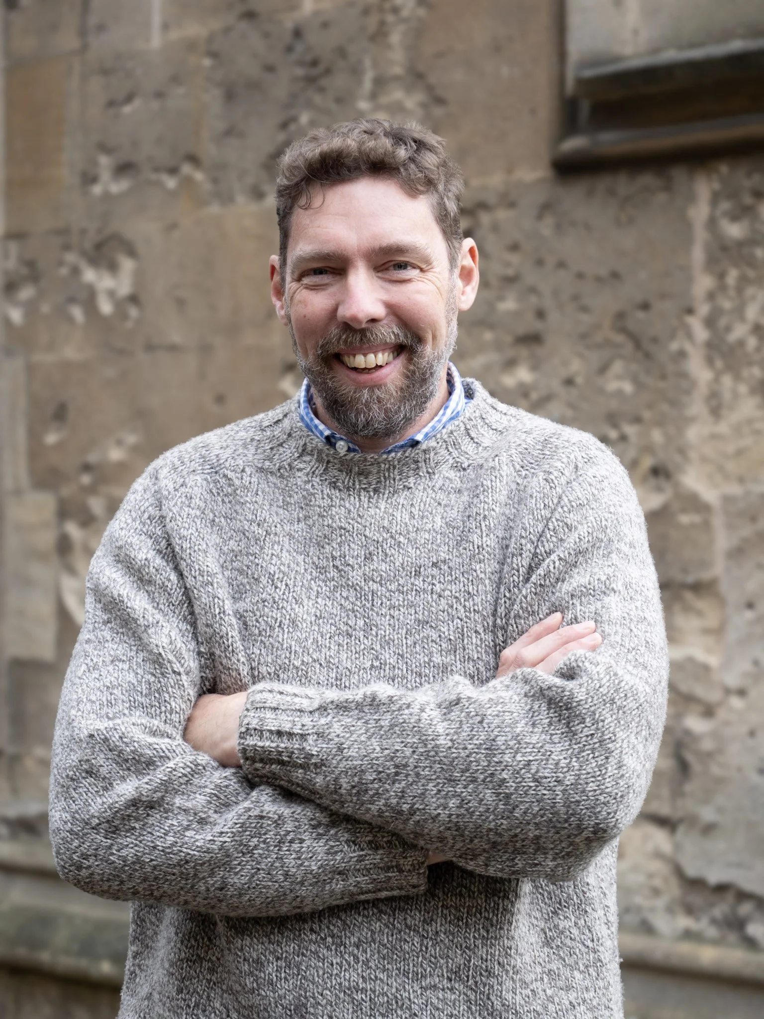 A man with a beard and curly hair, smiling with arms crossed, wearing a gray sweater over a blue and white checkered shirt, standing outdoors in front of a stone wall.