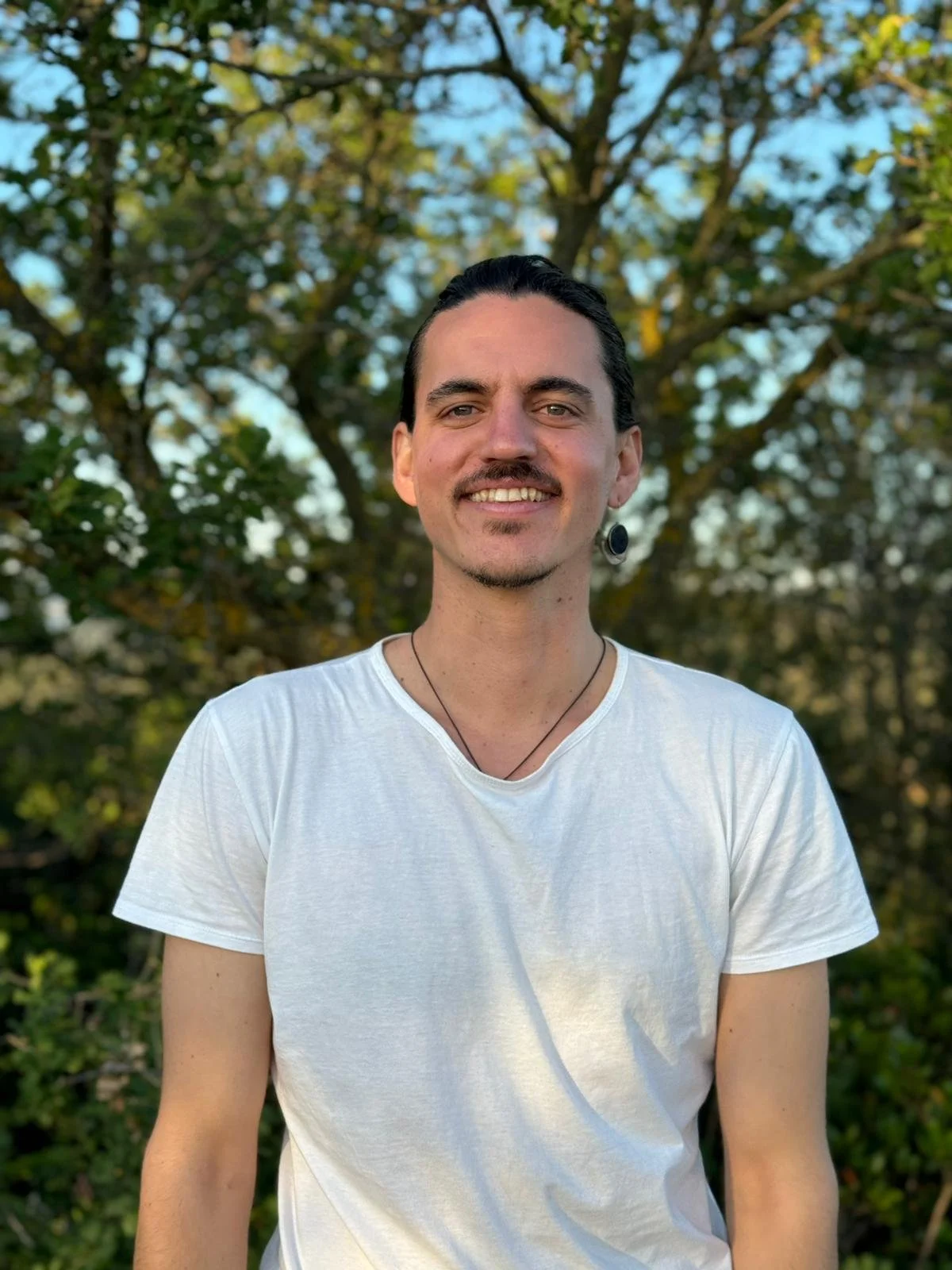 Portrait of a young man with dark hair, wearing a white T-shirt, smiling outdoors in front of trees with green leaves.