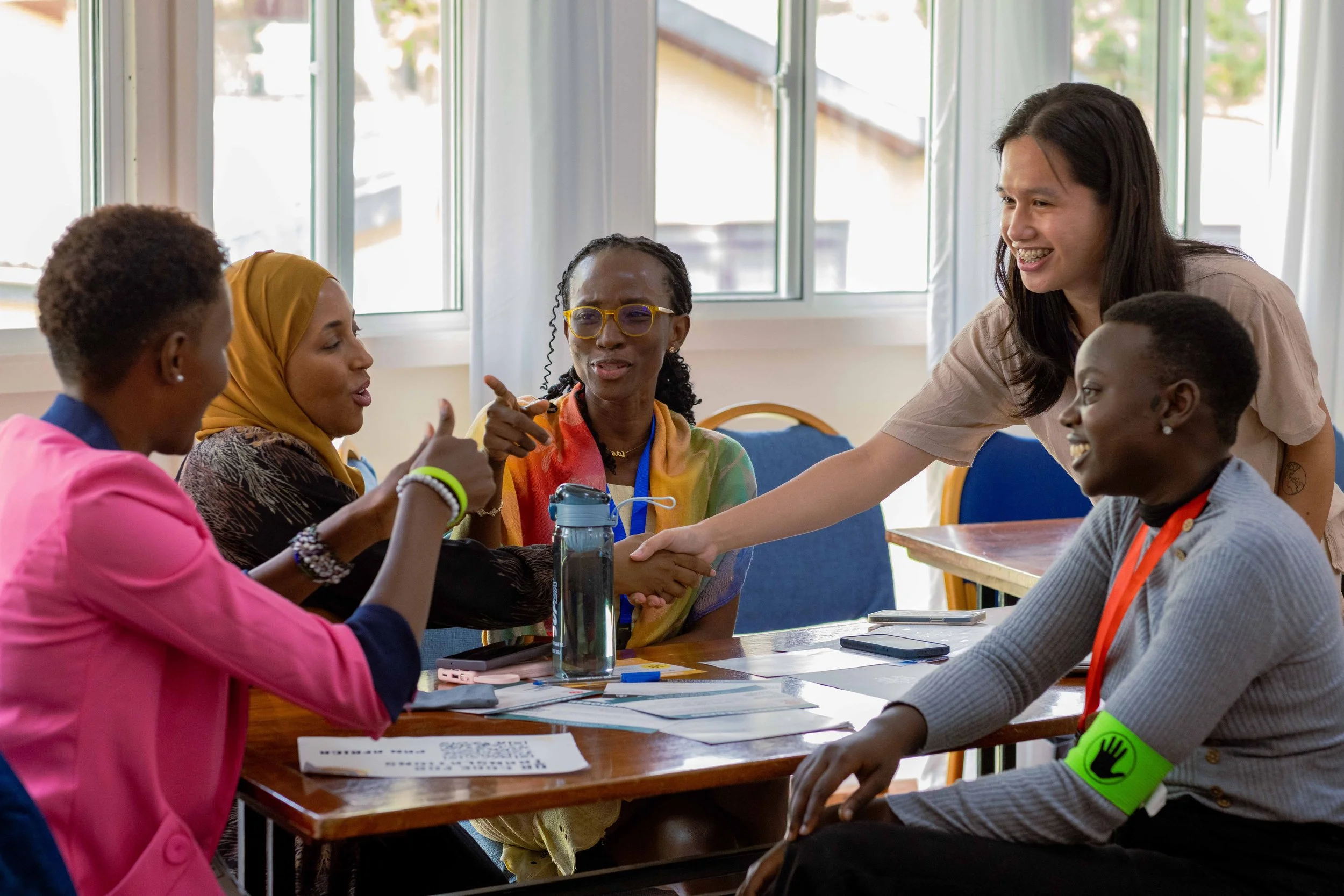 A diverse group of people gathered around a table in a bright room; one person is standing and shaking hands with a seated woman, both smiling. The others are engaged in conversation, with papers, a water bottle, and electronic devices on the table.