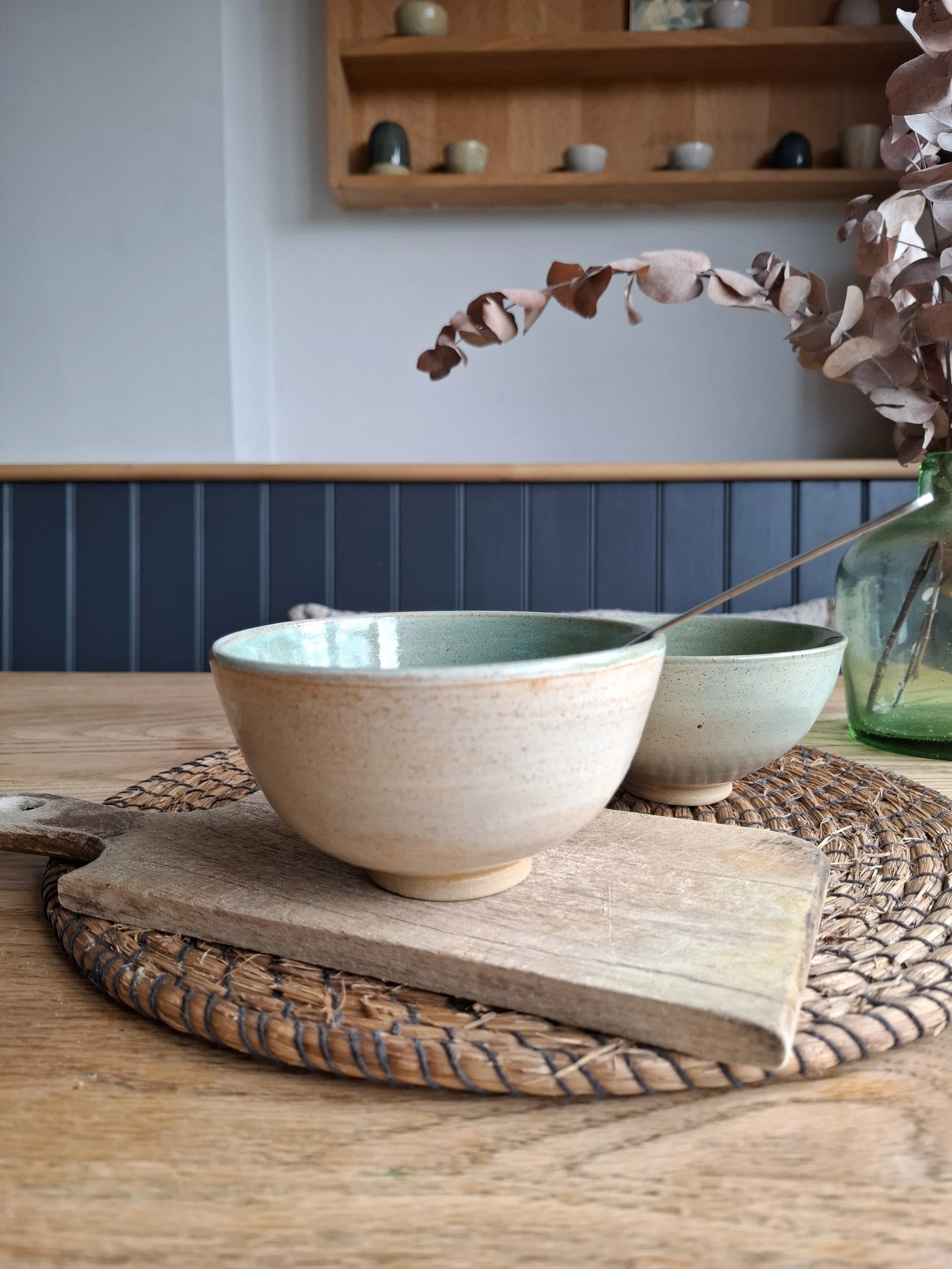 Two ceramic bowls on a small wooden board, placed on a woven placemat on a wooden table, with dried branches in a green glass vase in the background.