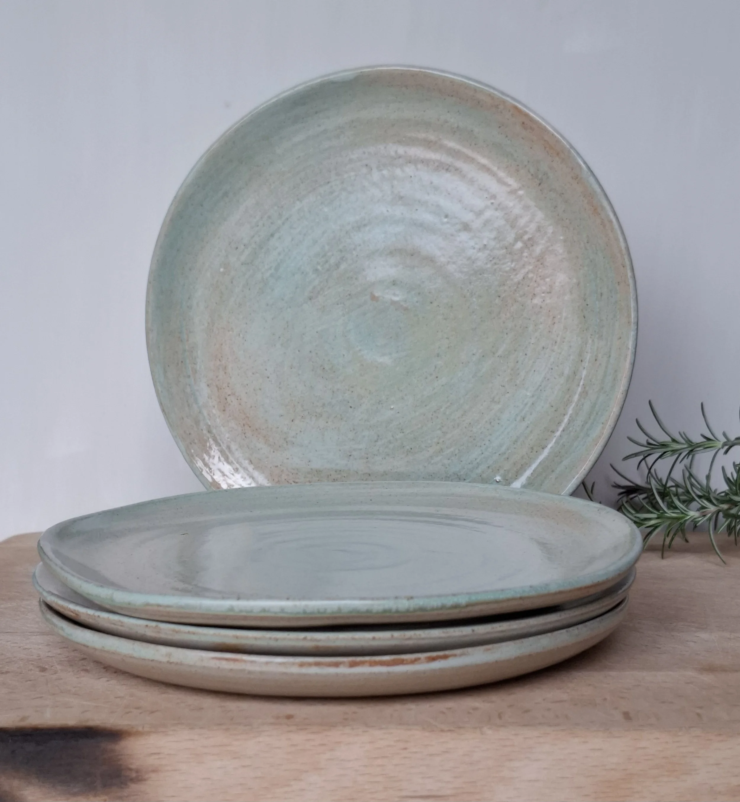 Set of five ceramic plates with a light, pastel-colored glaze, stacked on a wooden surface with a sprig of greenery beside them.