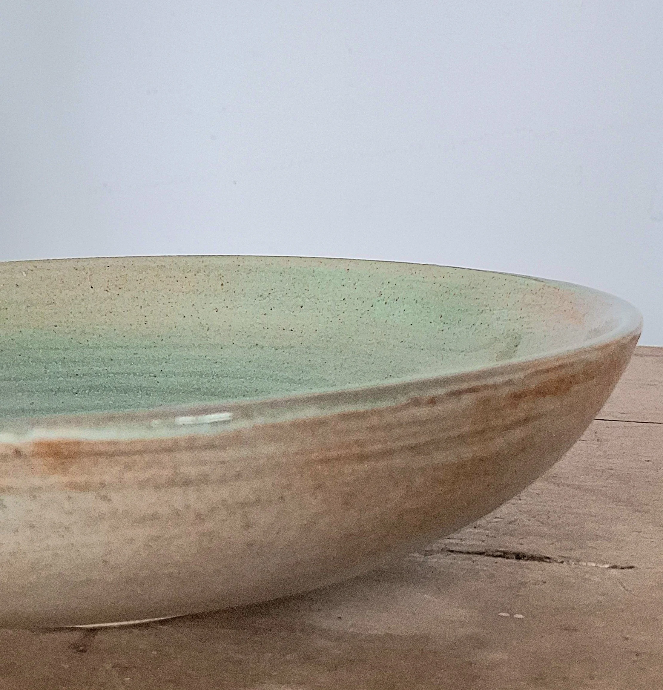 Close-up of a shallow ceramic bowl with a gradient glaze from light green to brown, placed on a wooden surface.
