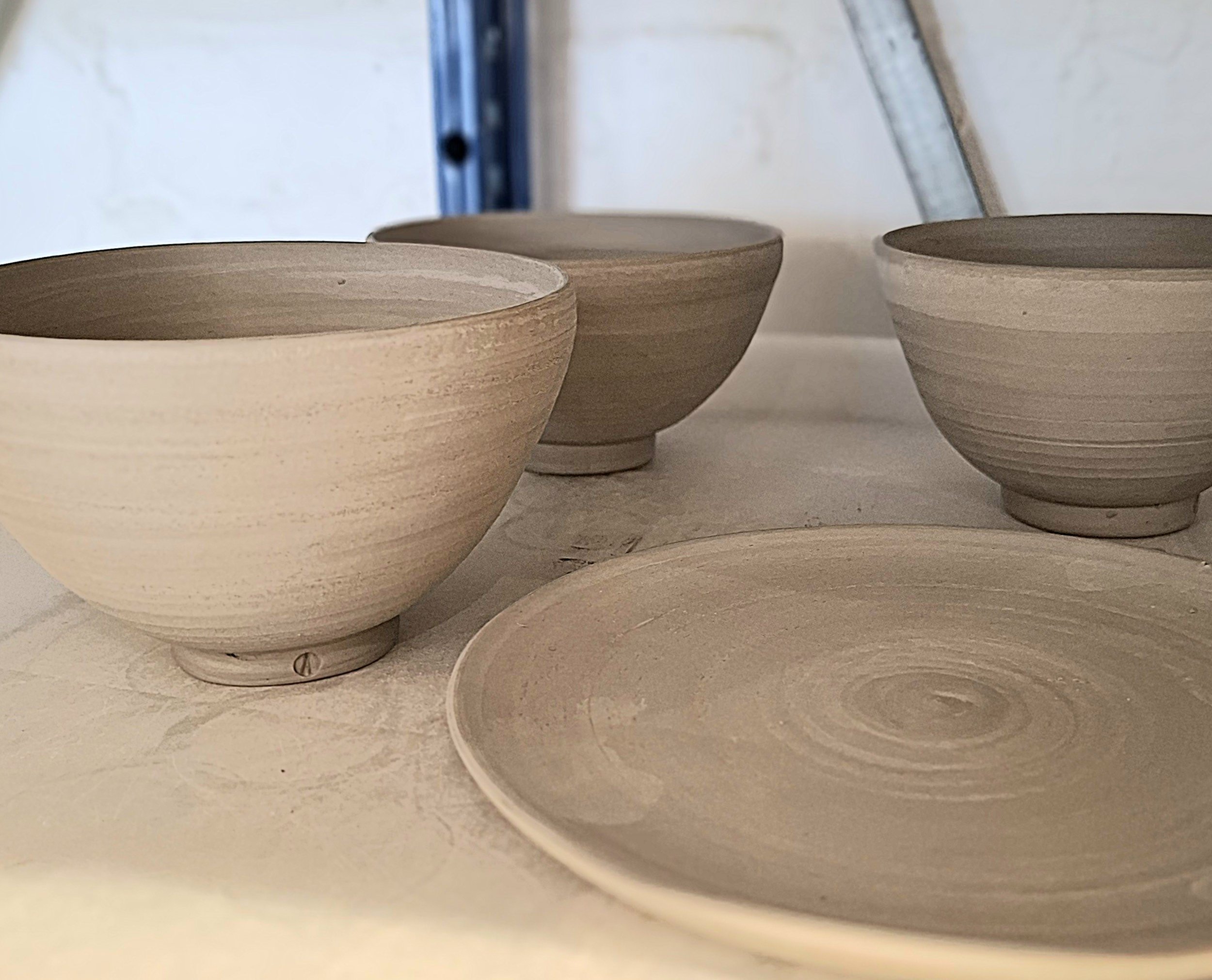 Three unfinished ceramic bowls and one round plate on a white shelf, with a metal ruler in the background.