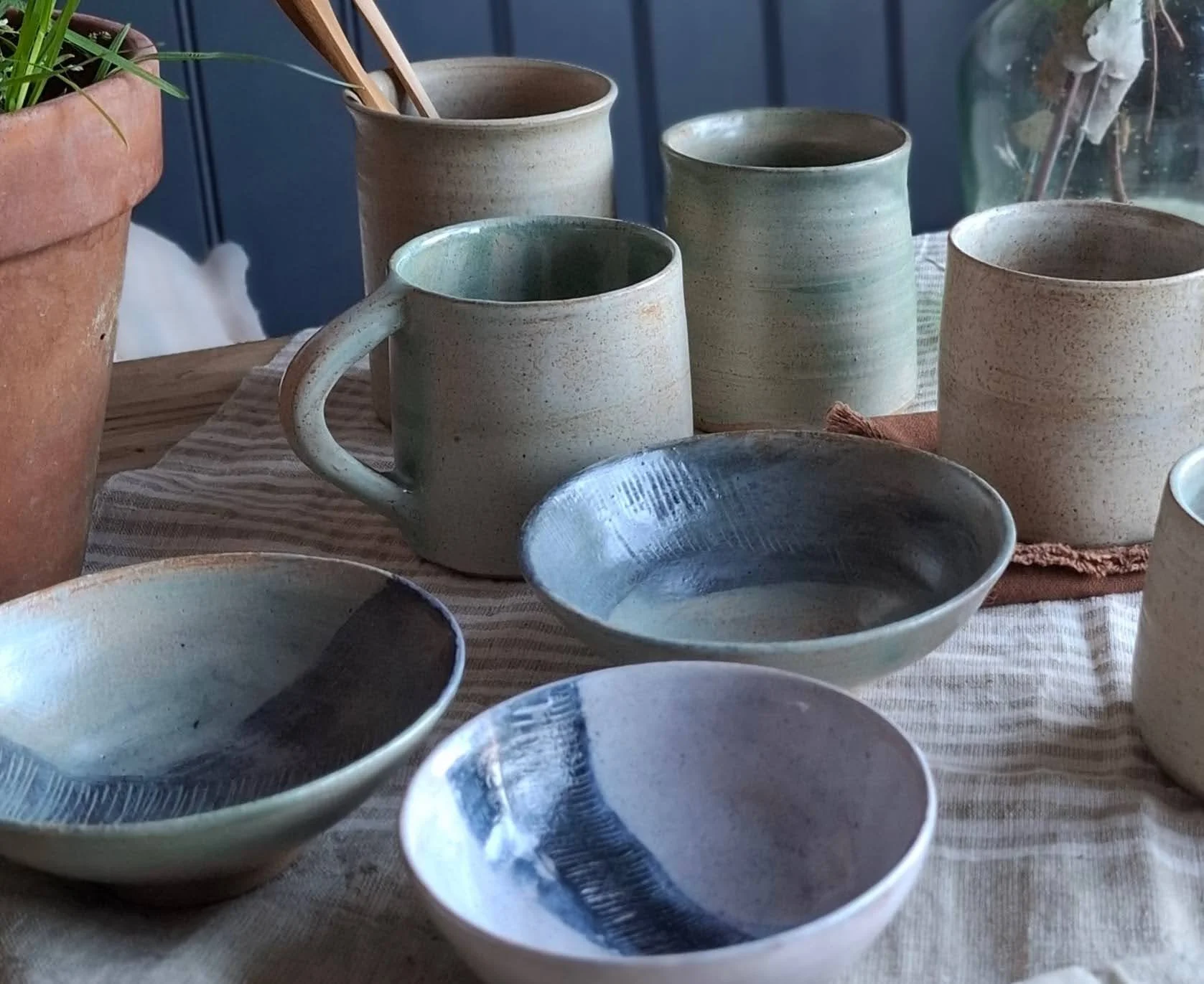 Several handmade ceramic bowls and mugs on a striped tablecloth, with a potted plant and a wooden background.