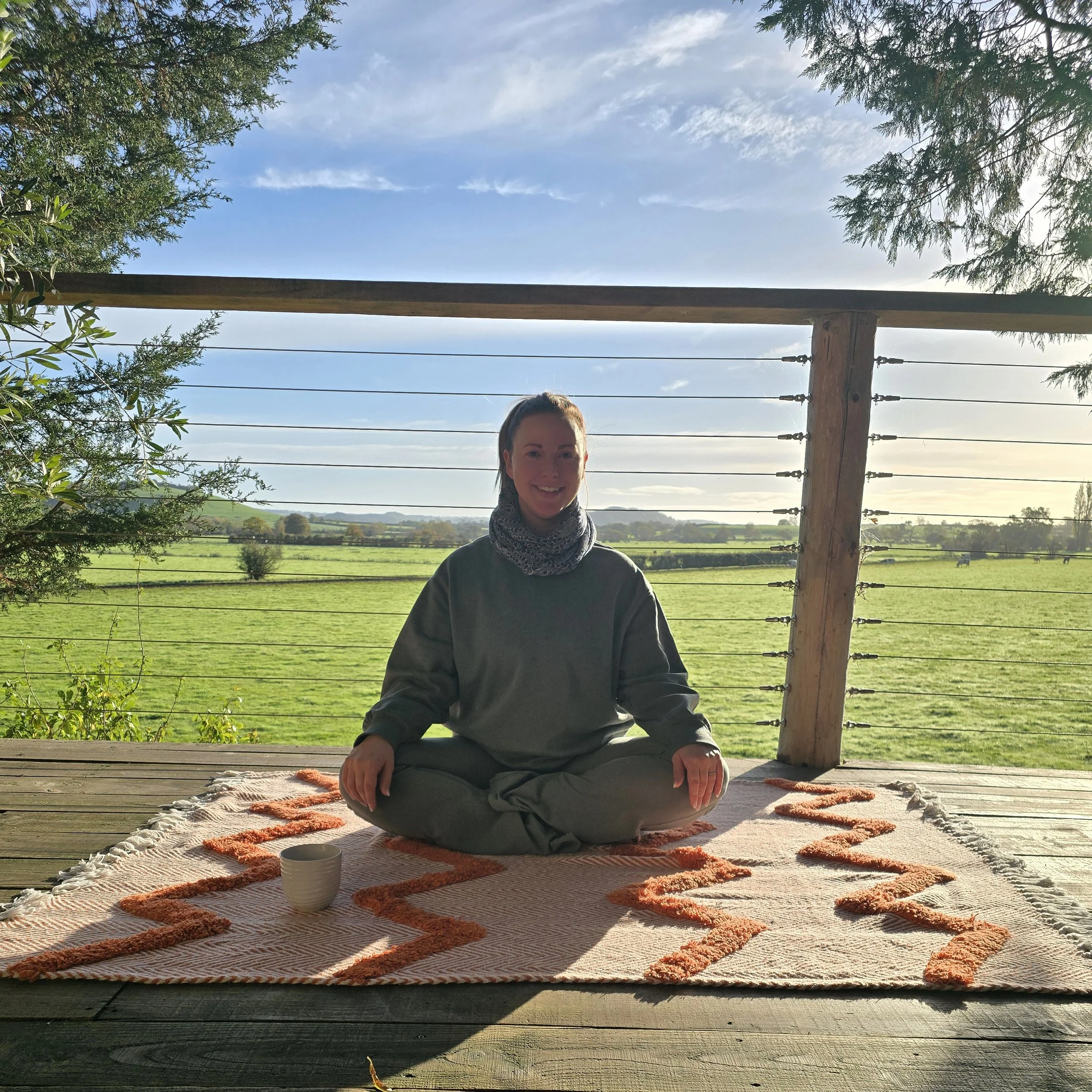 A woman practicing yoga outdoors on a colorful rug, with a scenic view of fields and trees in the background, under a wooden structure with ropes and trees around.