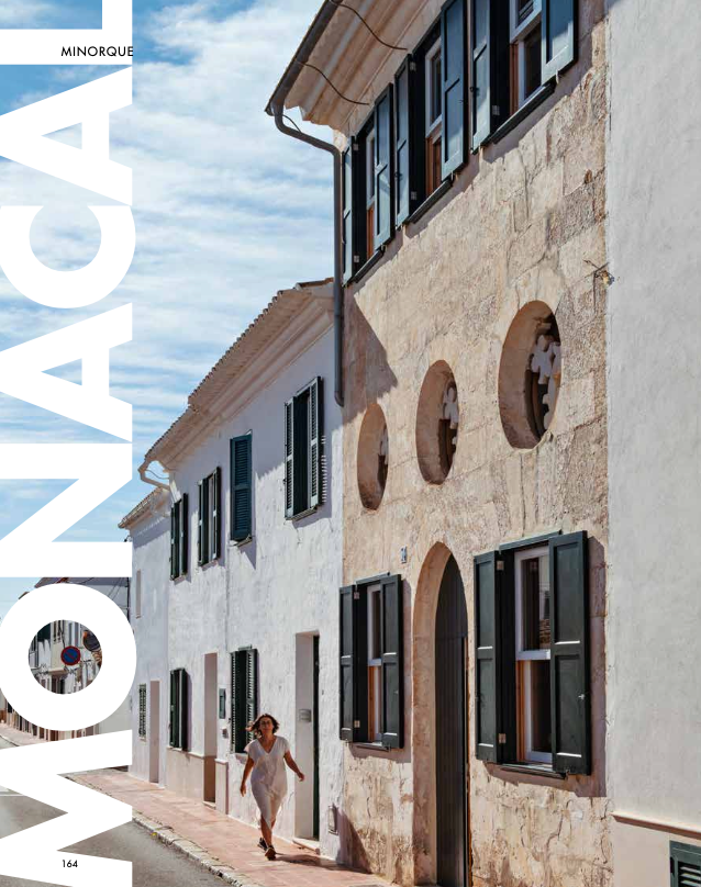 Casa antigua de piedra con ventanas y puertas de madera, en una calle soleada, con una mujer caminando.