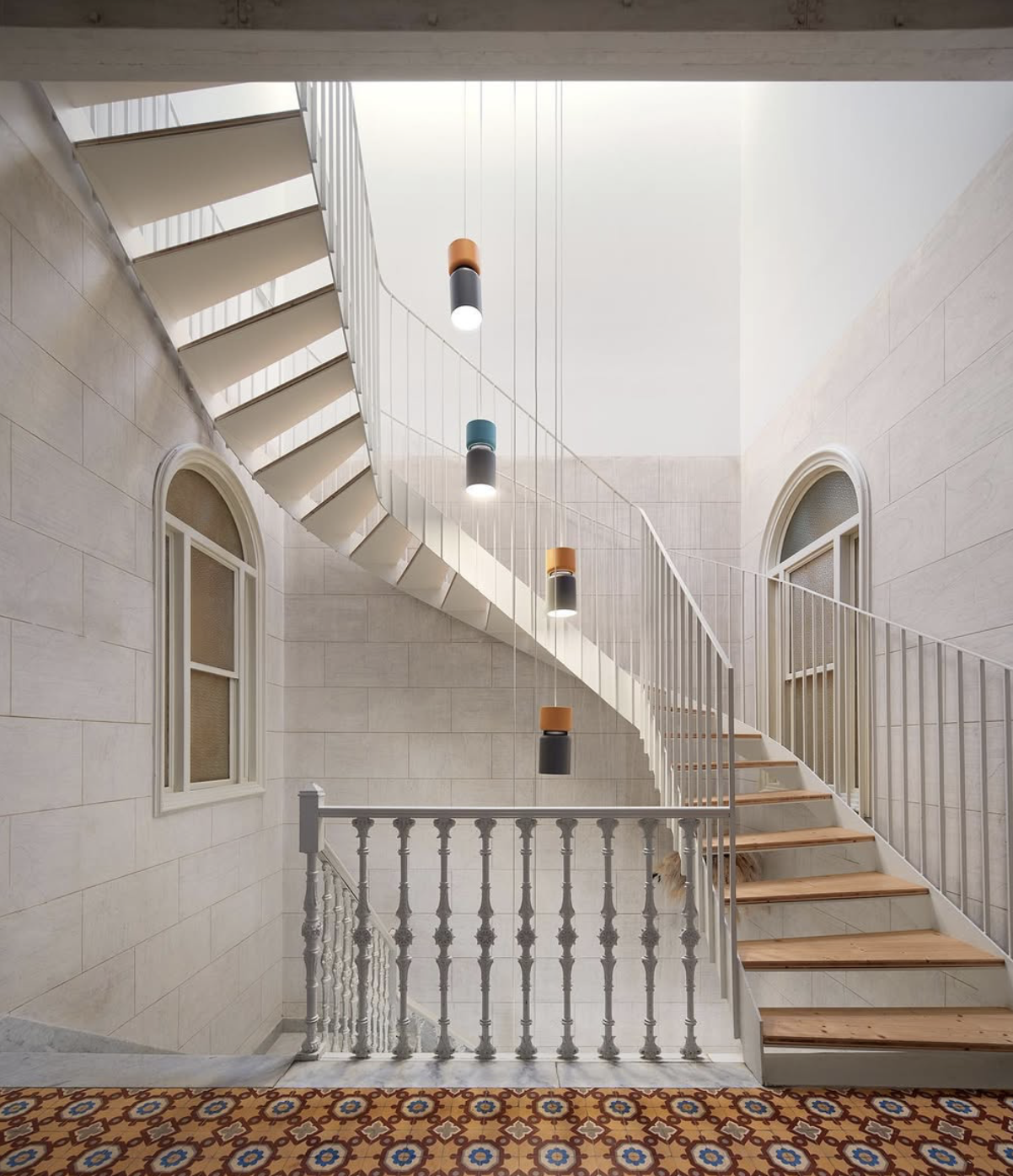 Escalera de caracol en un interior con paredes de piedra clara, ventanas arqueadas y lámparas colgantes de colores, con alfombra decorativa en el suelo.