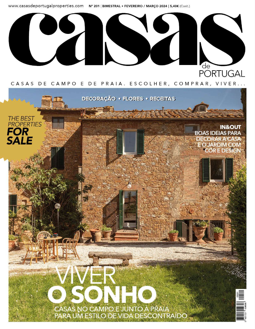 Una portada de revista que muestra una casa rústica de piedra en el campo, con ventanas con contraventanas verdes, un jardín con césped y muebles de exterior, y un cielo despejado.
