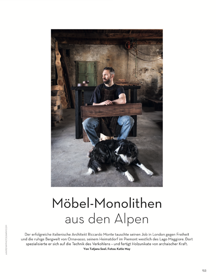 Un hombre con barba sentado en una silla en un taller con paredes de ladrillo y herramientas, acompañado de dos perros