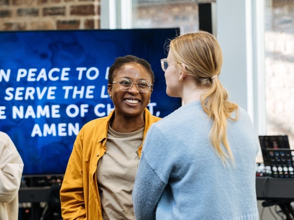 Two women discuss informally with a liturgical blessing displayed on a tv screen behind them
