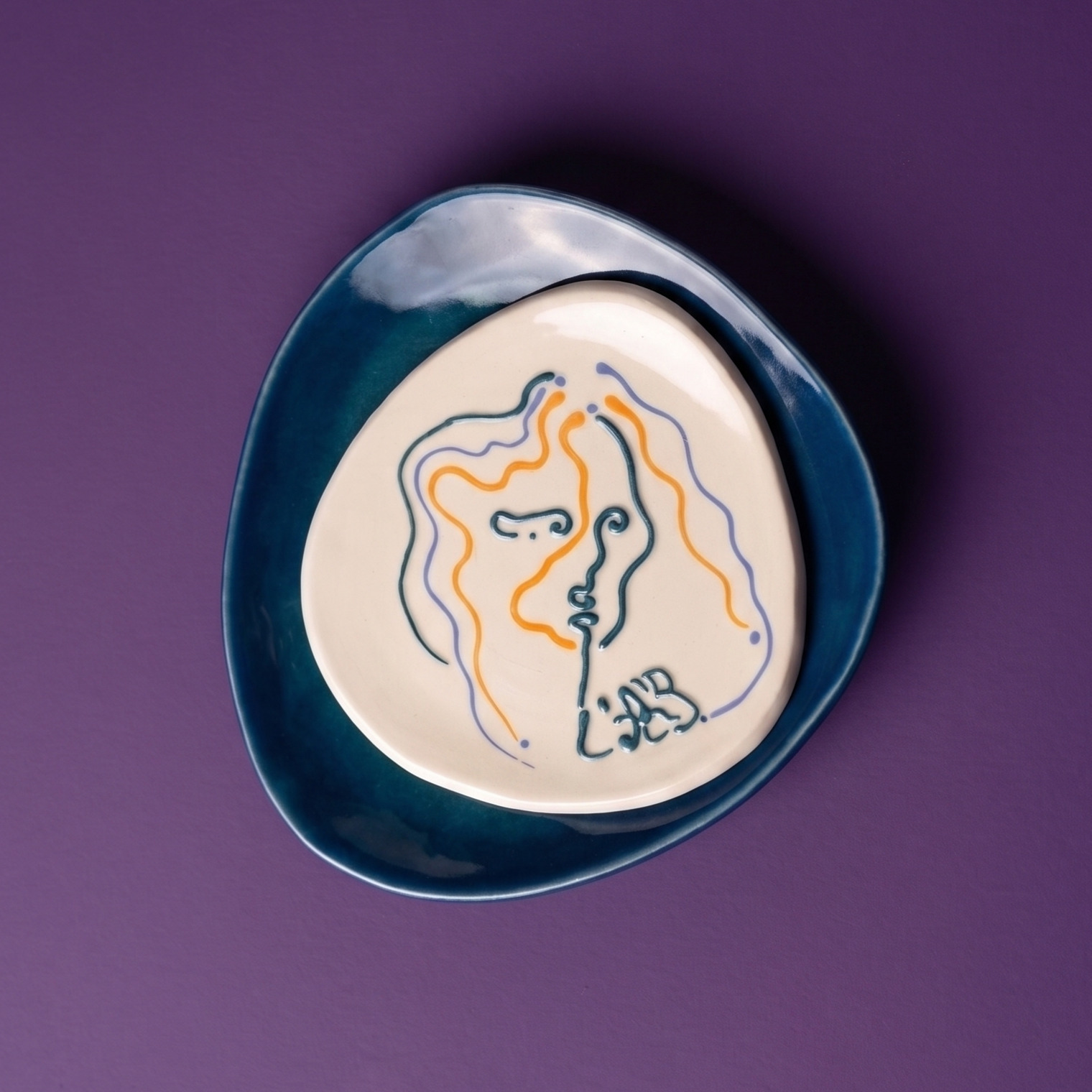 Assiette en céramique blanche avec un dessin coloré abstrait représentant une tête de profil et le mot « SPAR ». La plaque est posée sur une assiette plus grande bleu foncé, sur un fond violet.