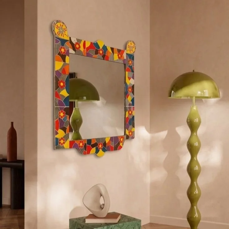 Un miroir coloré avec un cadre mosaïque, une lampe de sol verte avec un pied ondulé, et un mur beige dans un intérieur moderne.