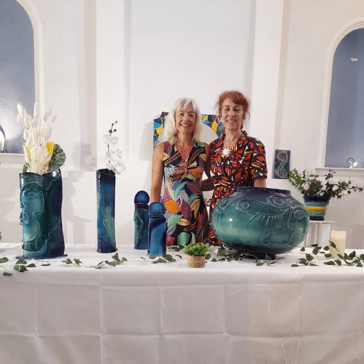 Deux femmes debout derrière une table ornée de vases en verre bleu avec des motifs, de fleurs et de plumes.