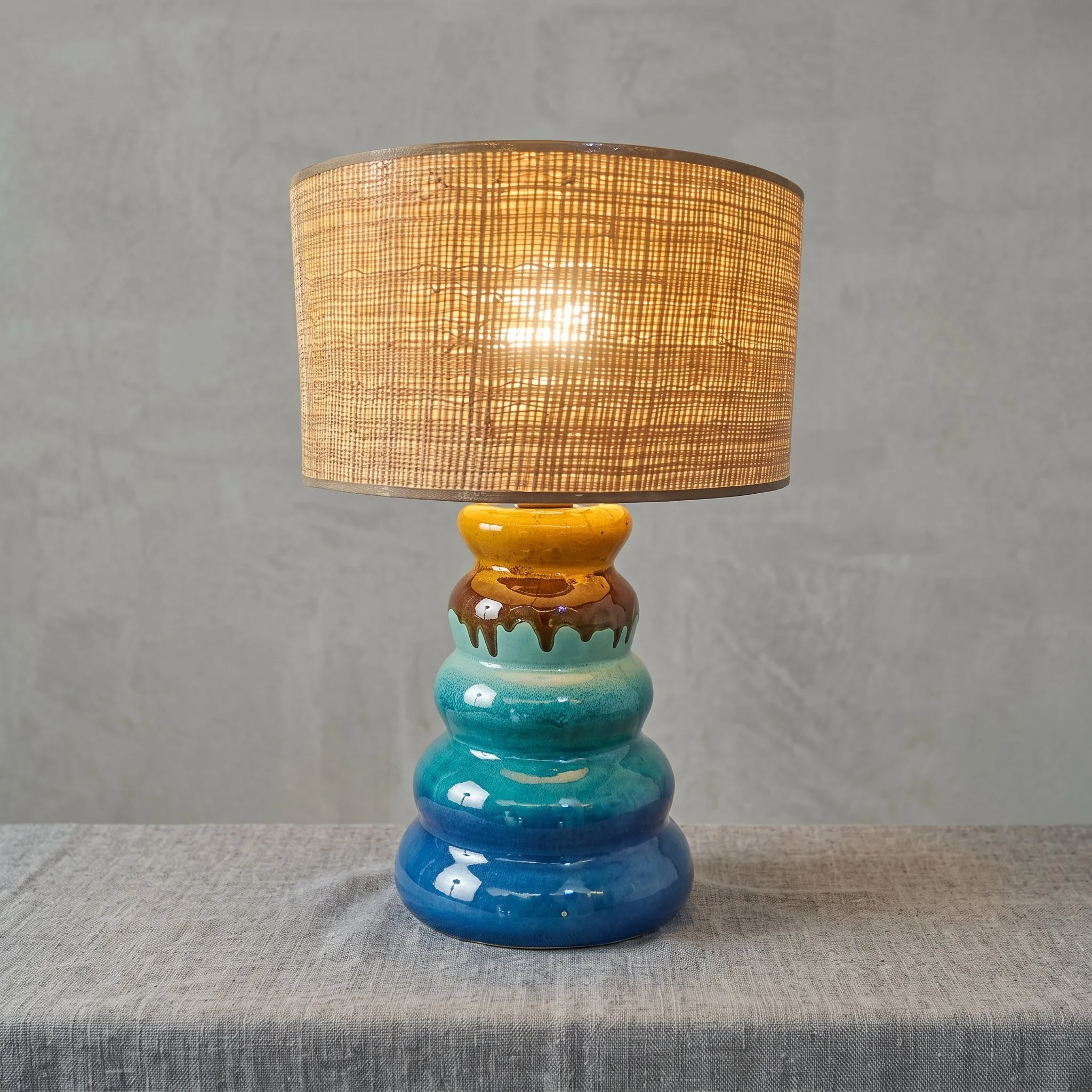 Une lampe de chevet avec un abat-jour en tissu beige lumineux et un socle en céramique colorée avec un motif de coulures de peinture, placée sur une surface en tissu gris à texture finie.