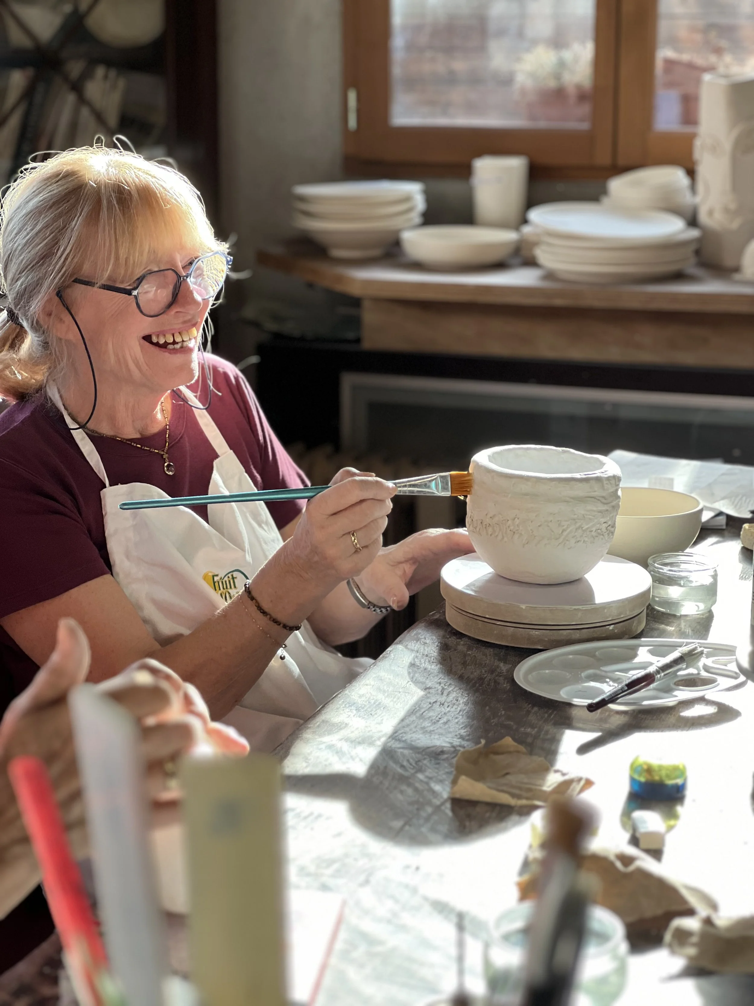 A woman with glasses and blonde hair painting details on a ceramic bowl in a pottery studio, smiling and enjoying her creative activity.