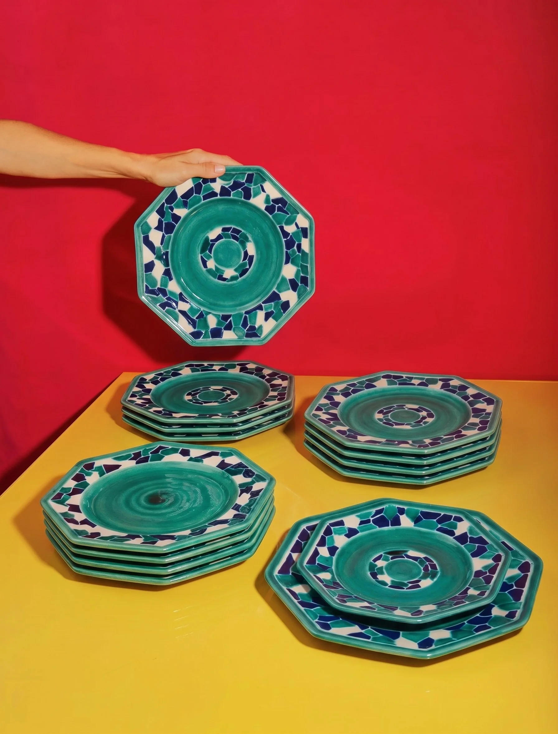 Une main tient un plateau en céramique vert et bleu avec un motif mosaïque, entouré de plusieurs autres plateaux empilés sur une table jaune, contre un mur rouge.